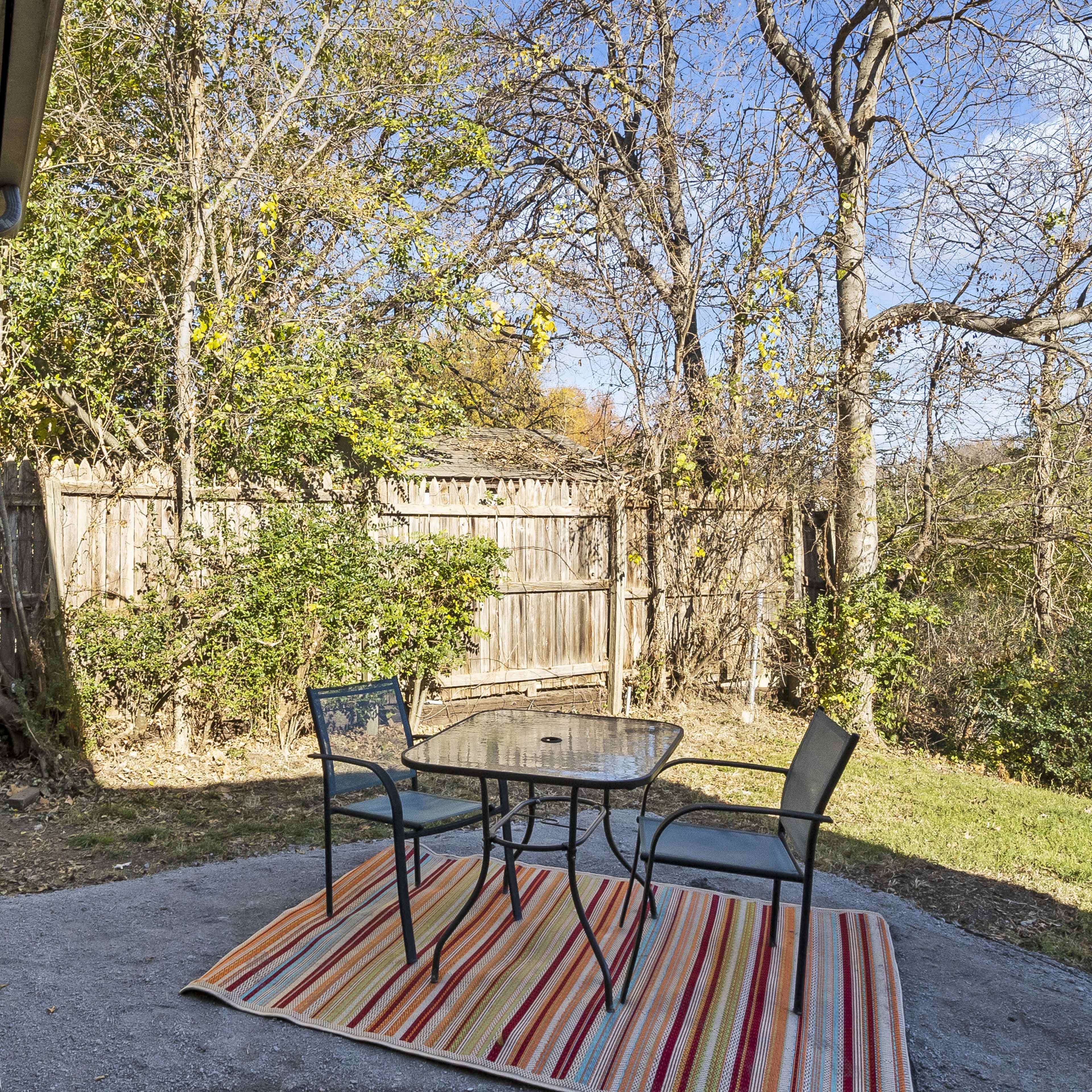 A patio area features a glass table with four black chairs on a striped rug, surrounded by trees and a wooden fence.