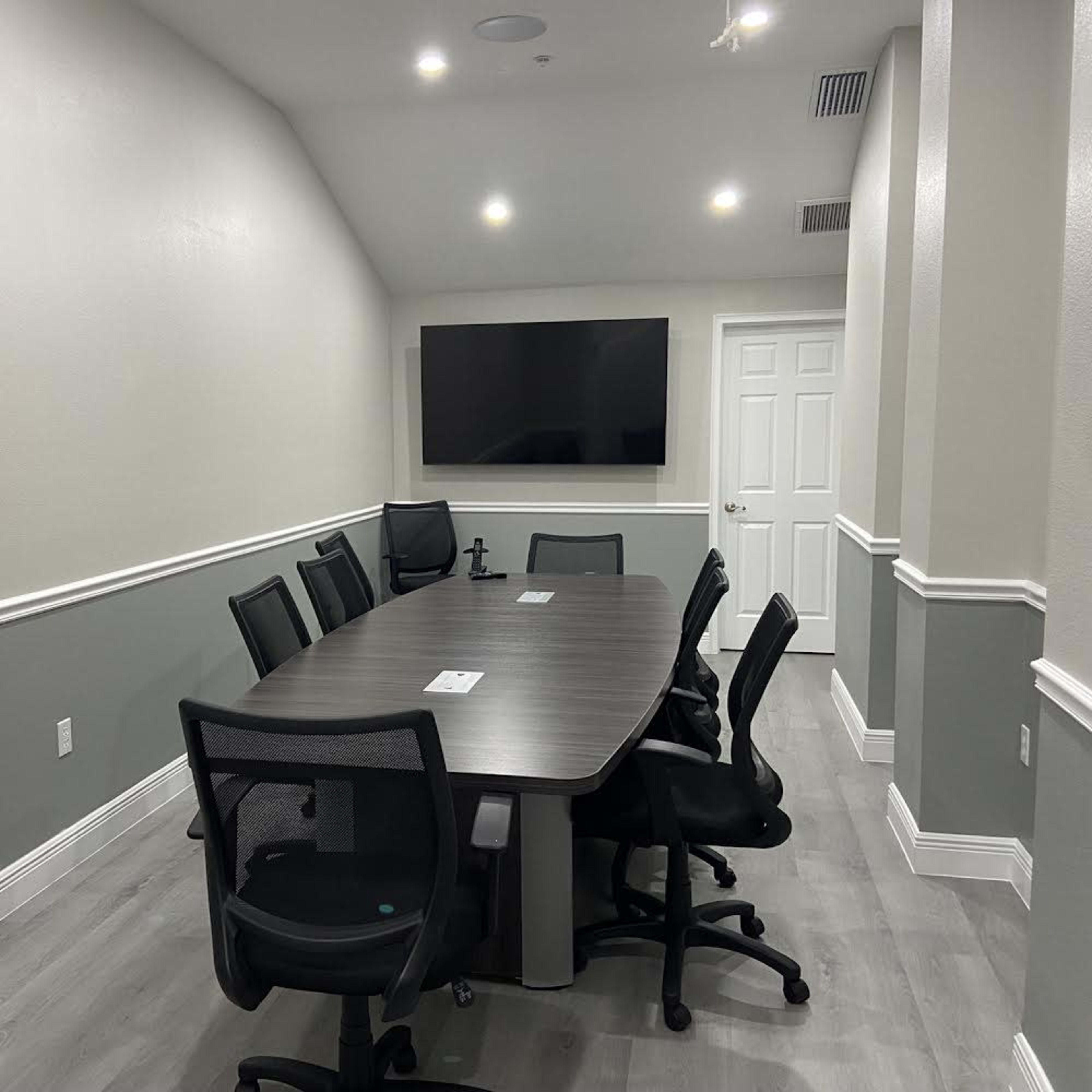 A modern conference room with a long table surrounded by eight office chairs, a wall-mounted screen, and a door at the far end.