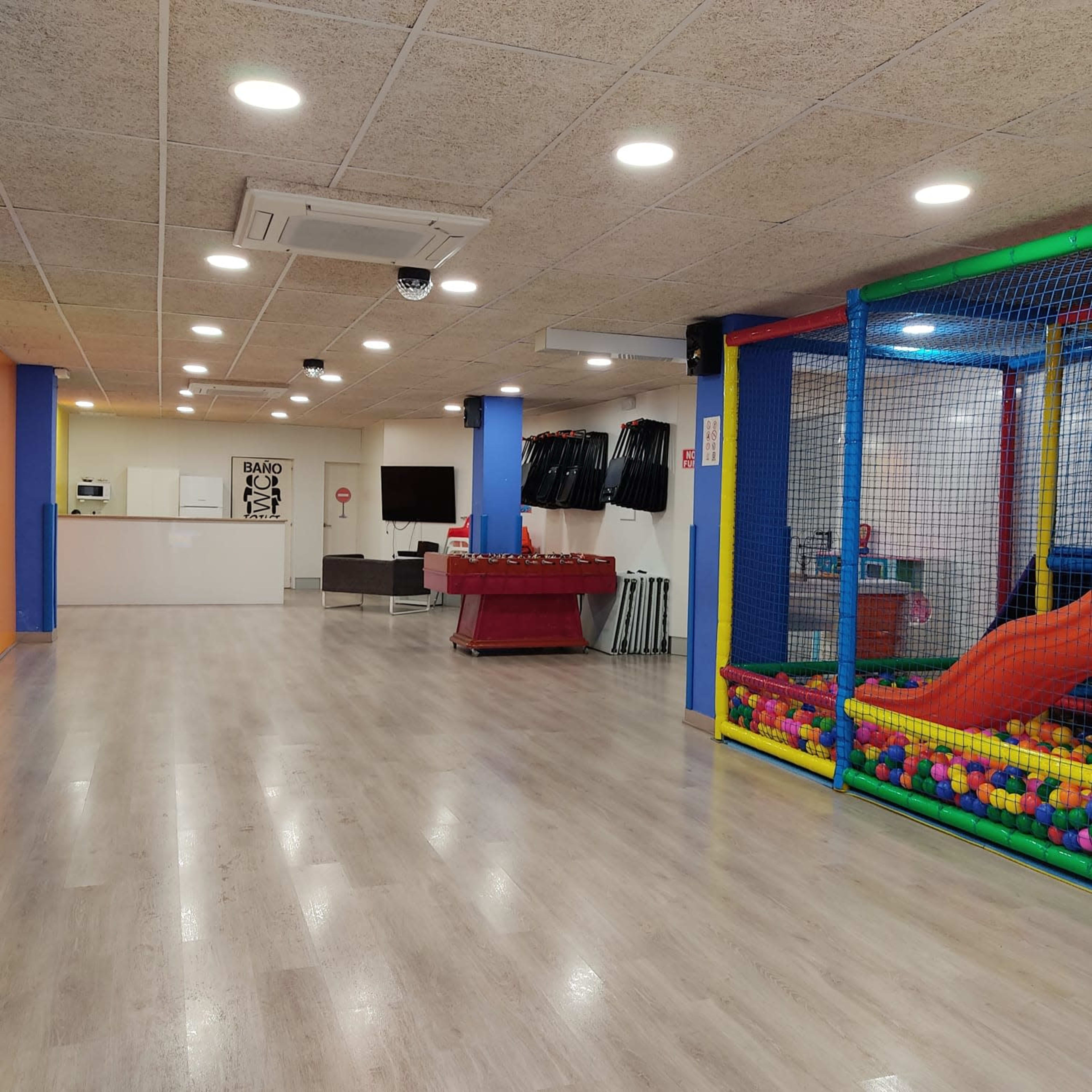 A spacious indoor play area featuring a soft play structure with slides and colorful balls on one side, and a kitchenette and seating area on the opposite side.