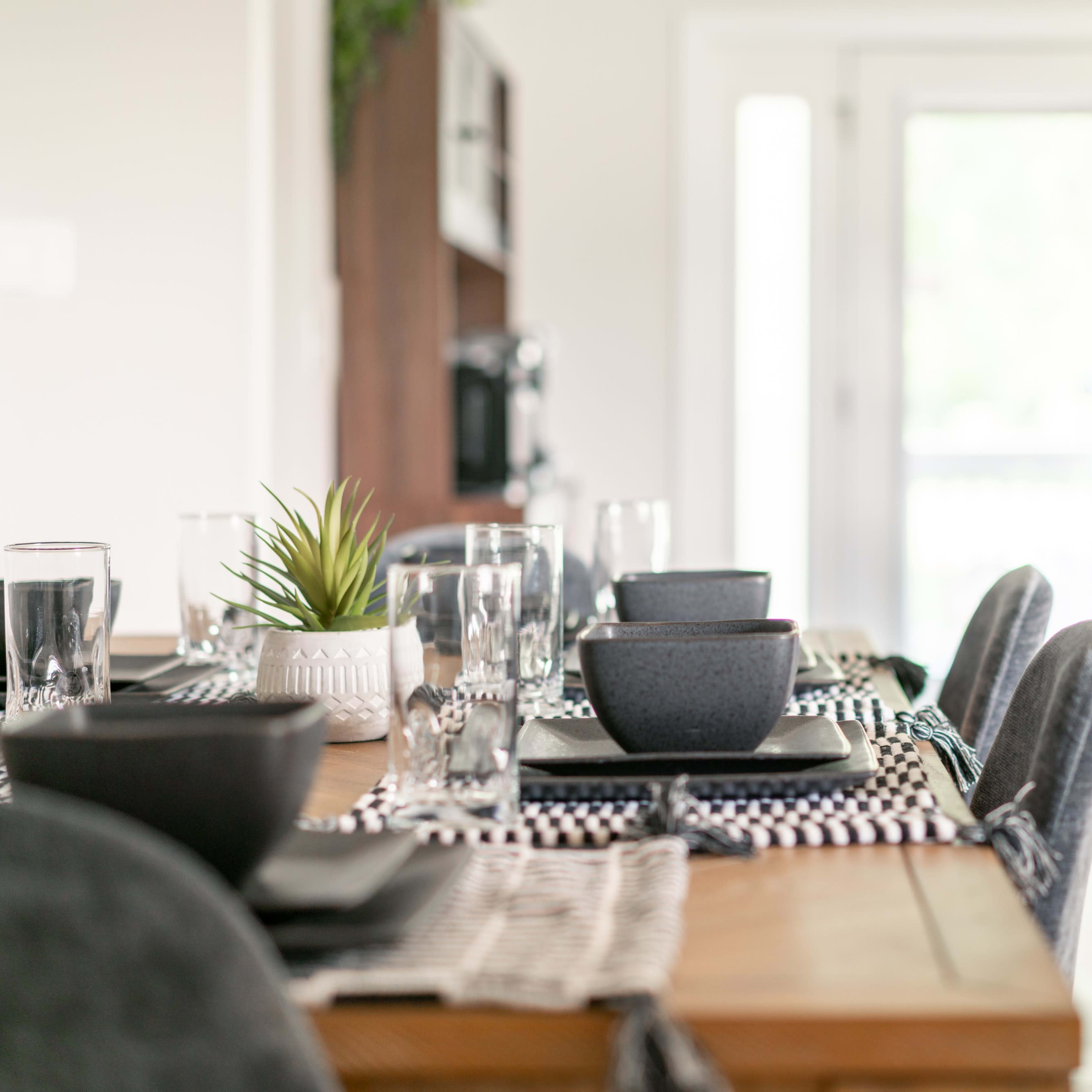 A dining table is set with black bowls, glasses of water, and decorative plants, with a view of a modern kitchen in the background.
