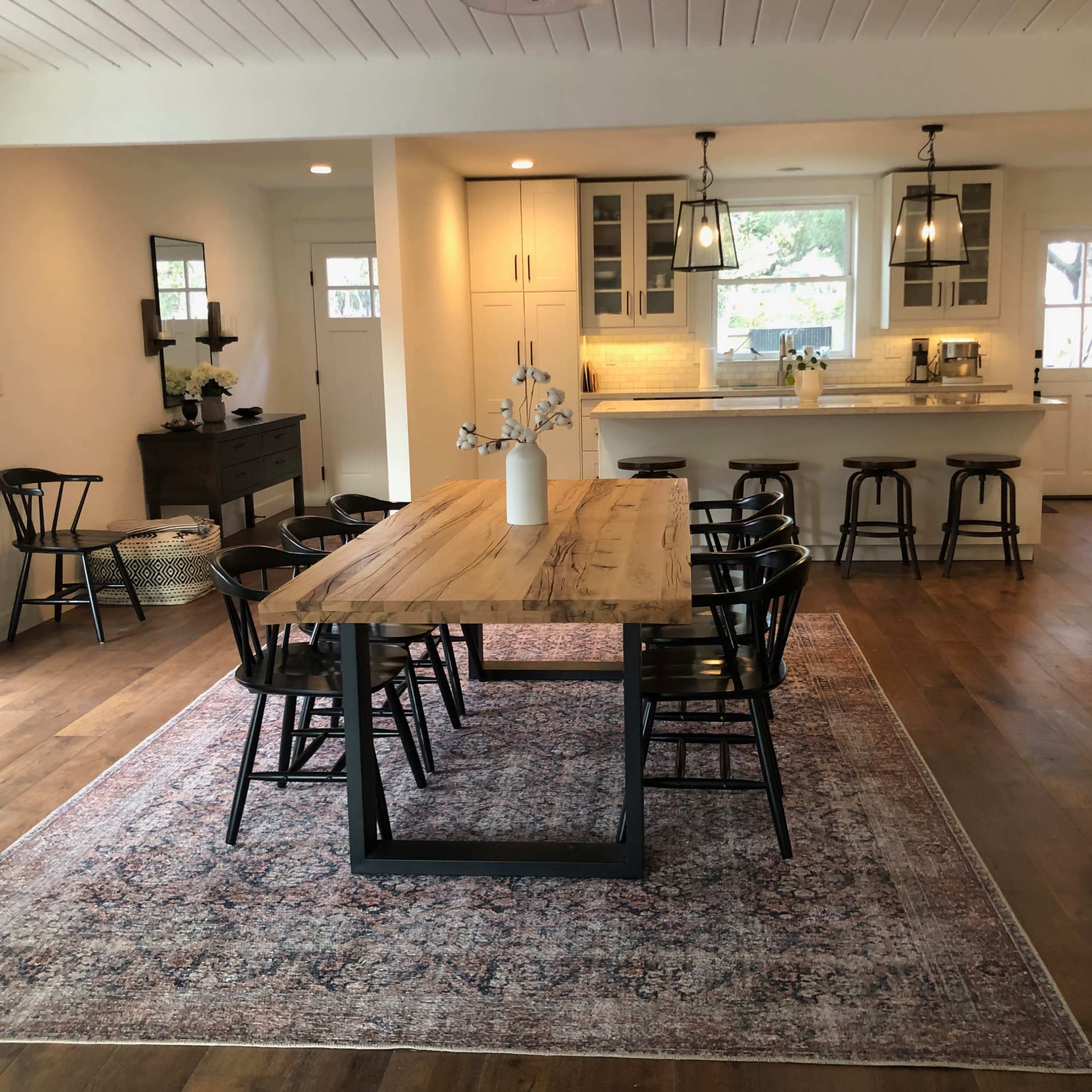 A dining area with a large wooden table surrounded by black chairs, set on a patterned rug, adjacent to a kitchen featuring light cabinetry and pendant lighting.