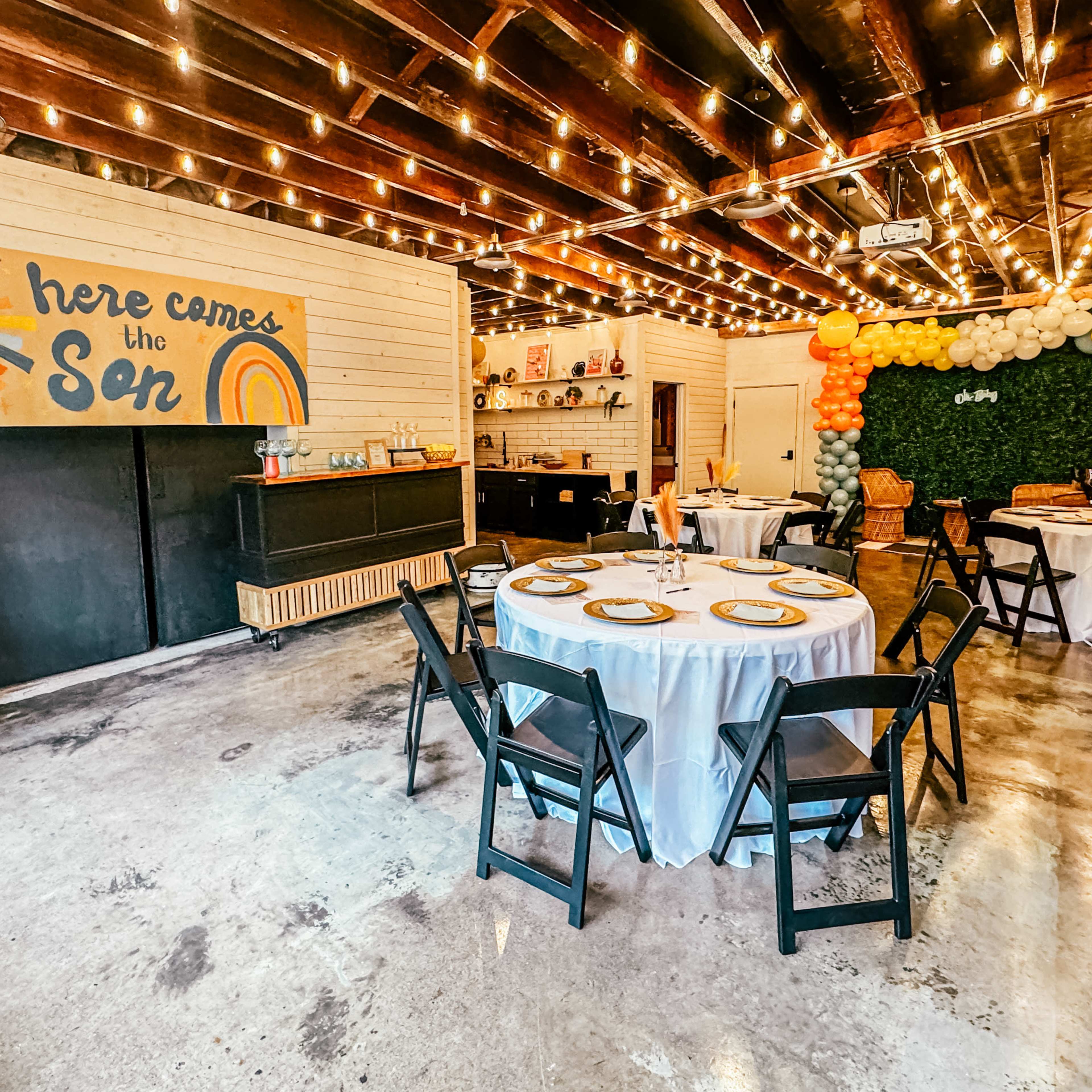 A decorated event space with round tables set up, a colorful mural on the wall, and strings of lights hanging from the ceiling.