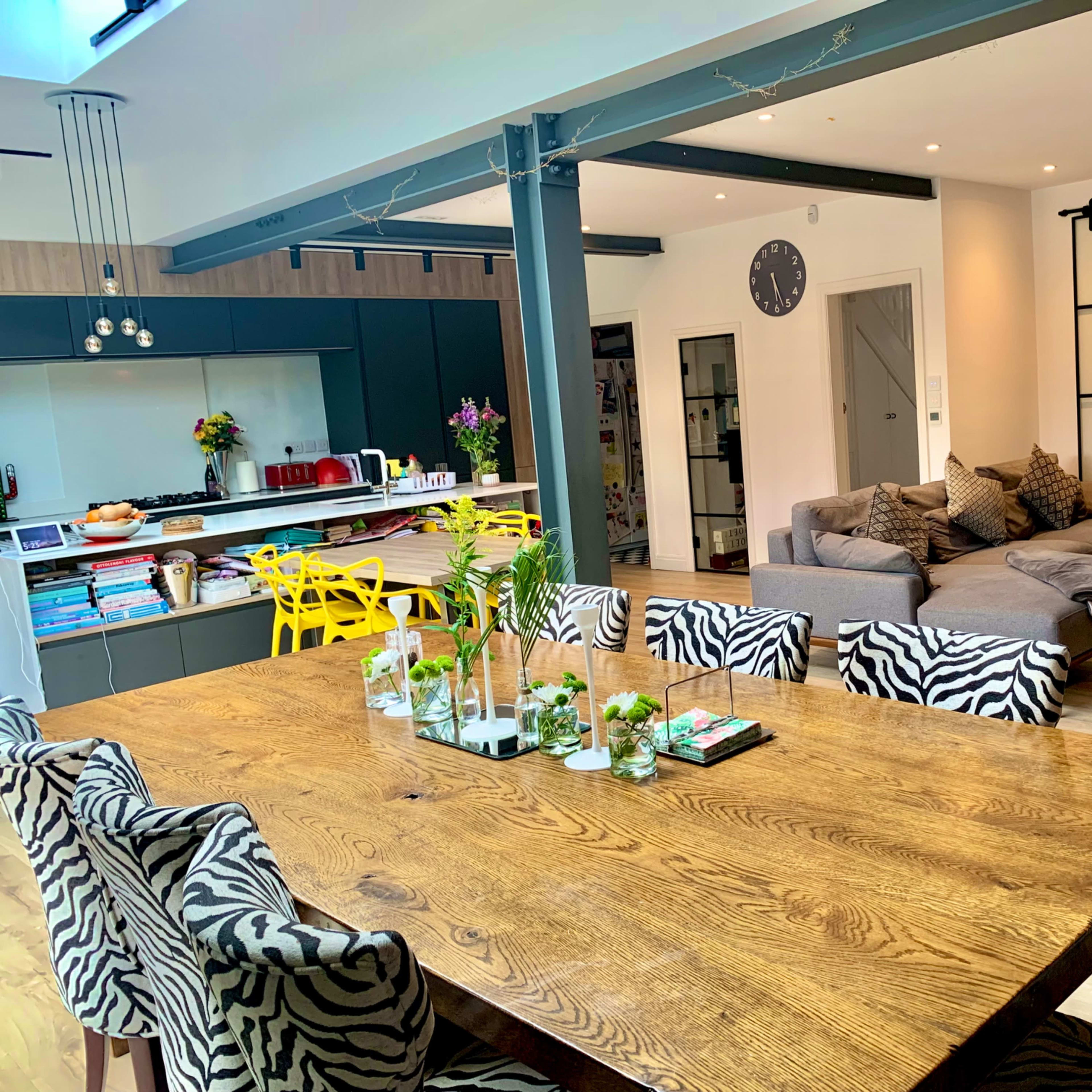 A modern open-plan living space featuring a large wooden dining table with zebra-print chairs, a sleek kitchen area with a breakfast bar, and a cozy seating area with a sectional couch.