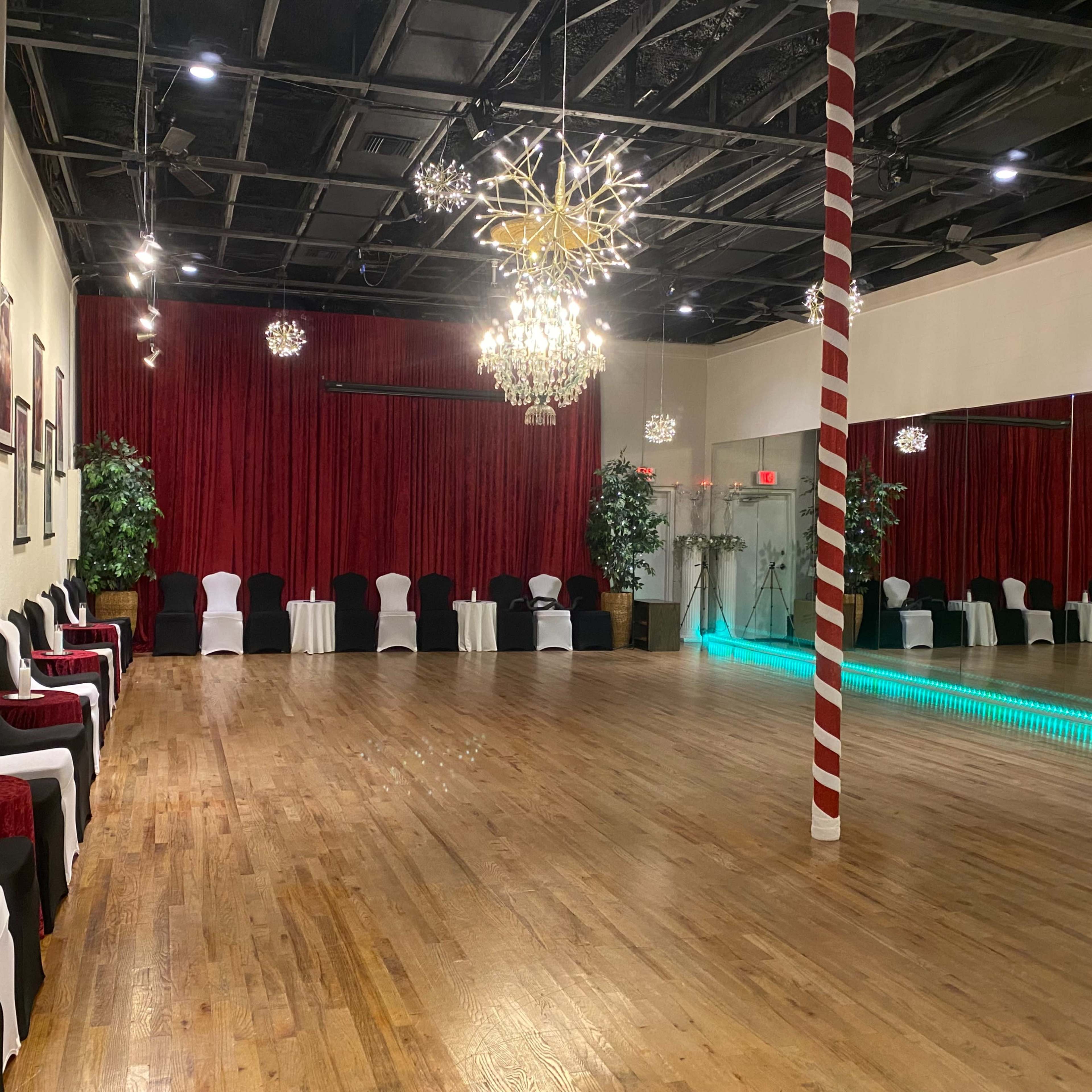 A spacious dance studio with a wooden floor, red curtains, and decorative lighting, including chandeliers and a row of chairs along the walls.