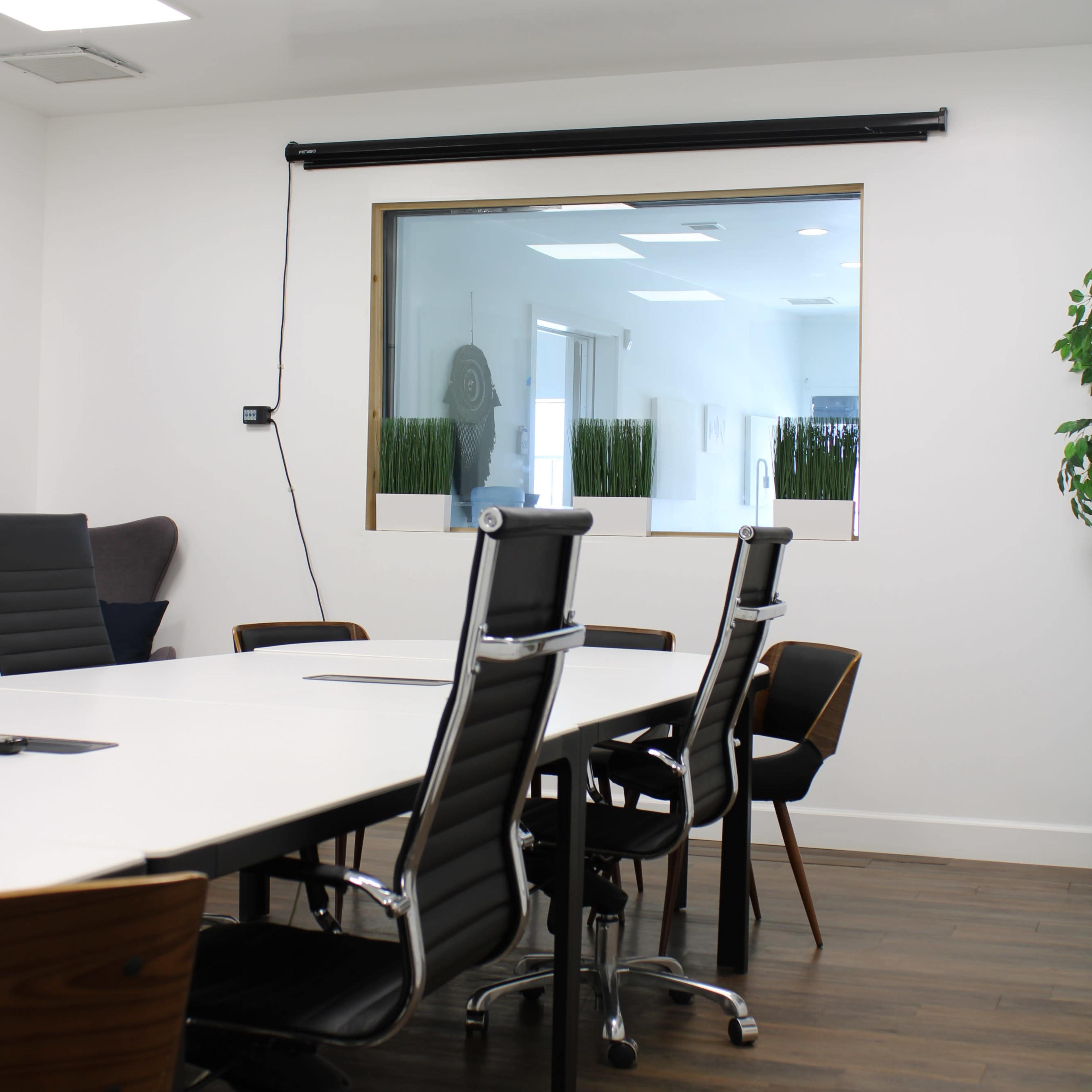 A modern conference room features a large table surrounded by black and wooden chairs, with a window overlooking a bright, plant-filled area.