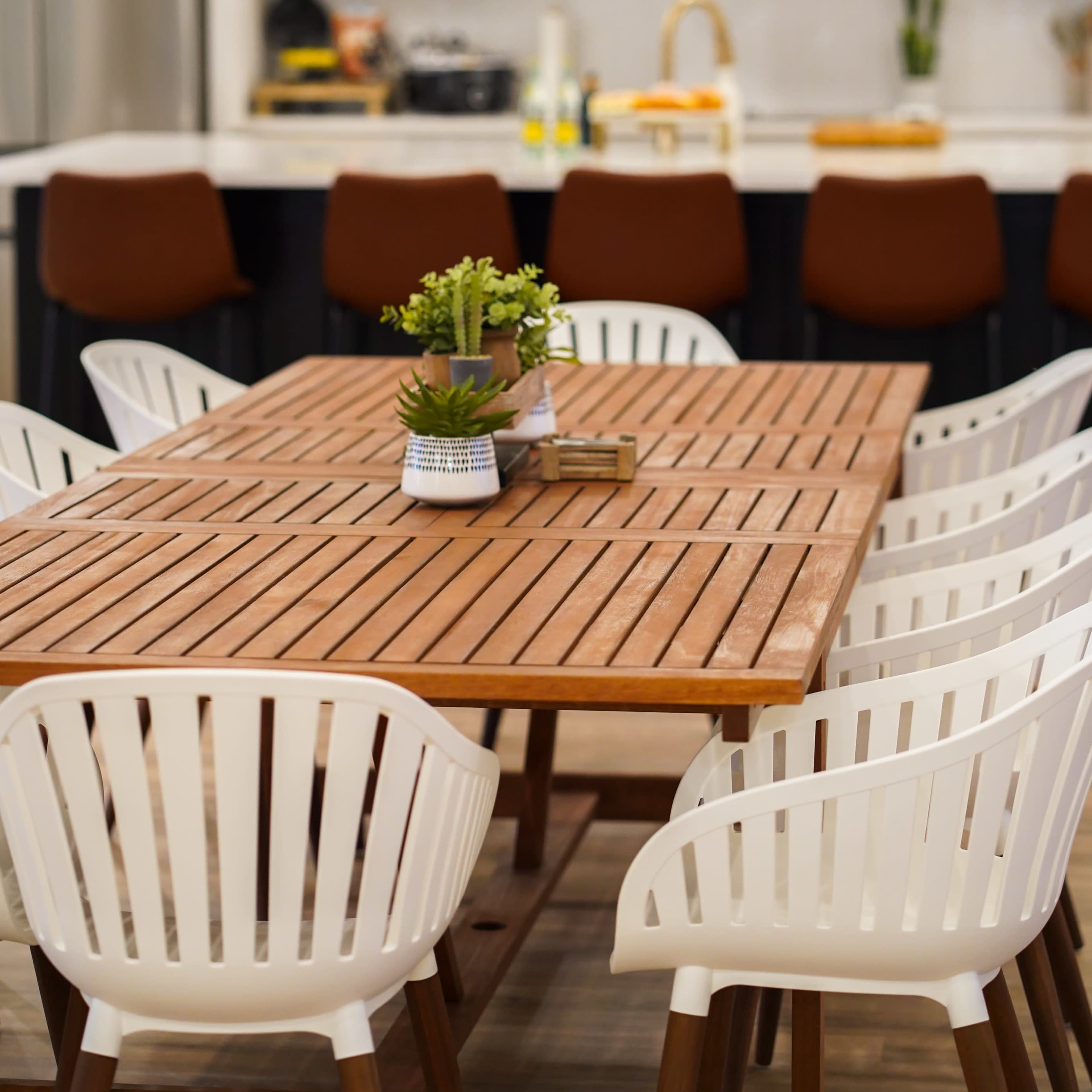 A wooden dining table surrounded by white chairs is positioned in a modern kitchen with a large island and brown bar stools.
