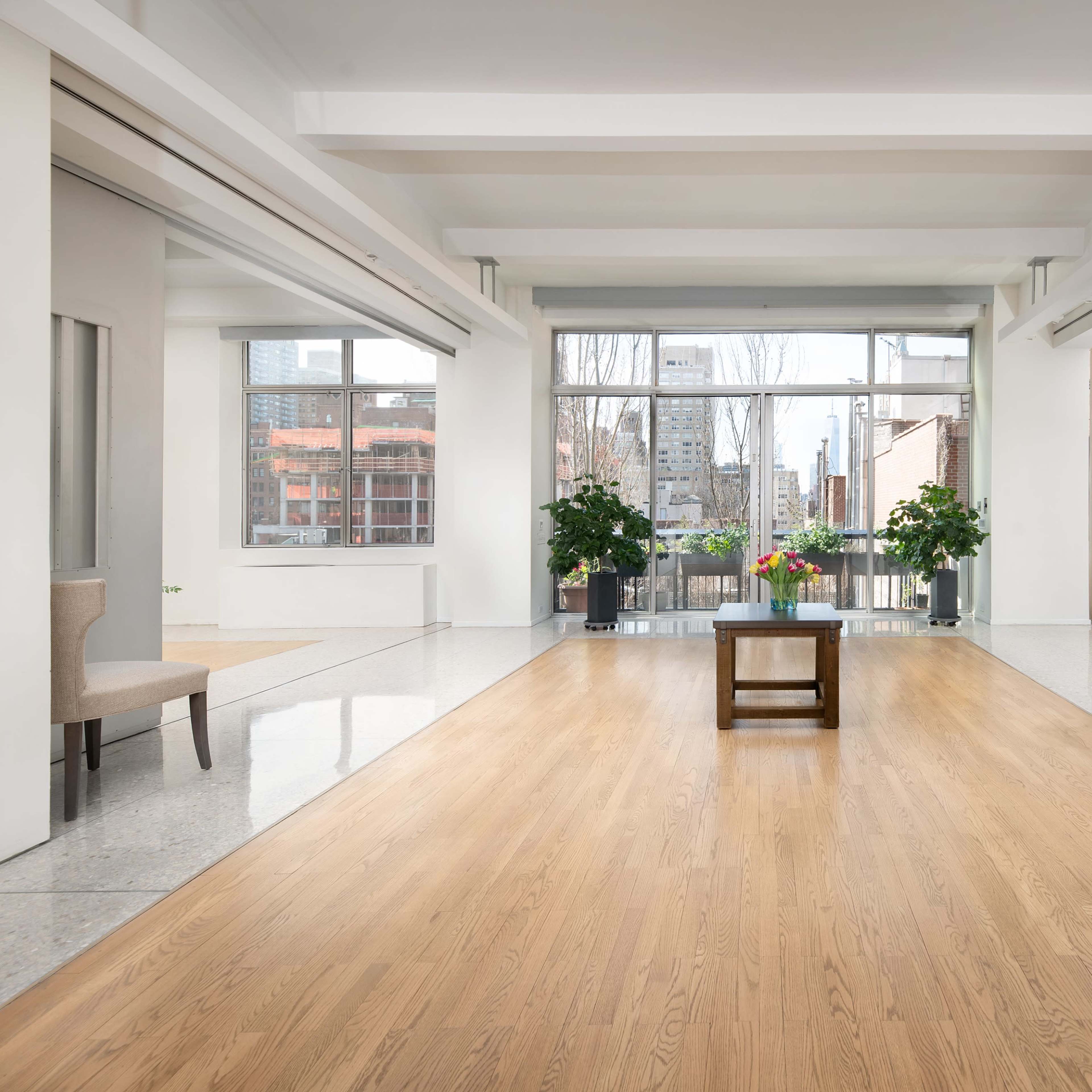 A spacious interior features hardwood flooring, two chairs on either side, and a central wooden table with potted plants, framed by large windows displaying a city view.