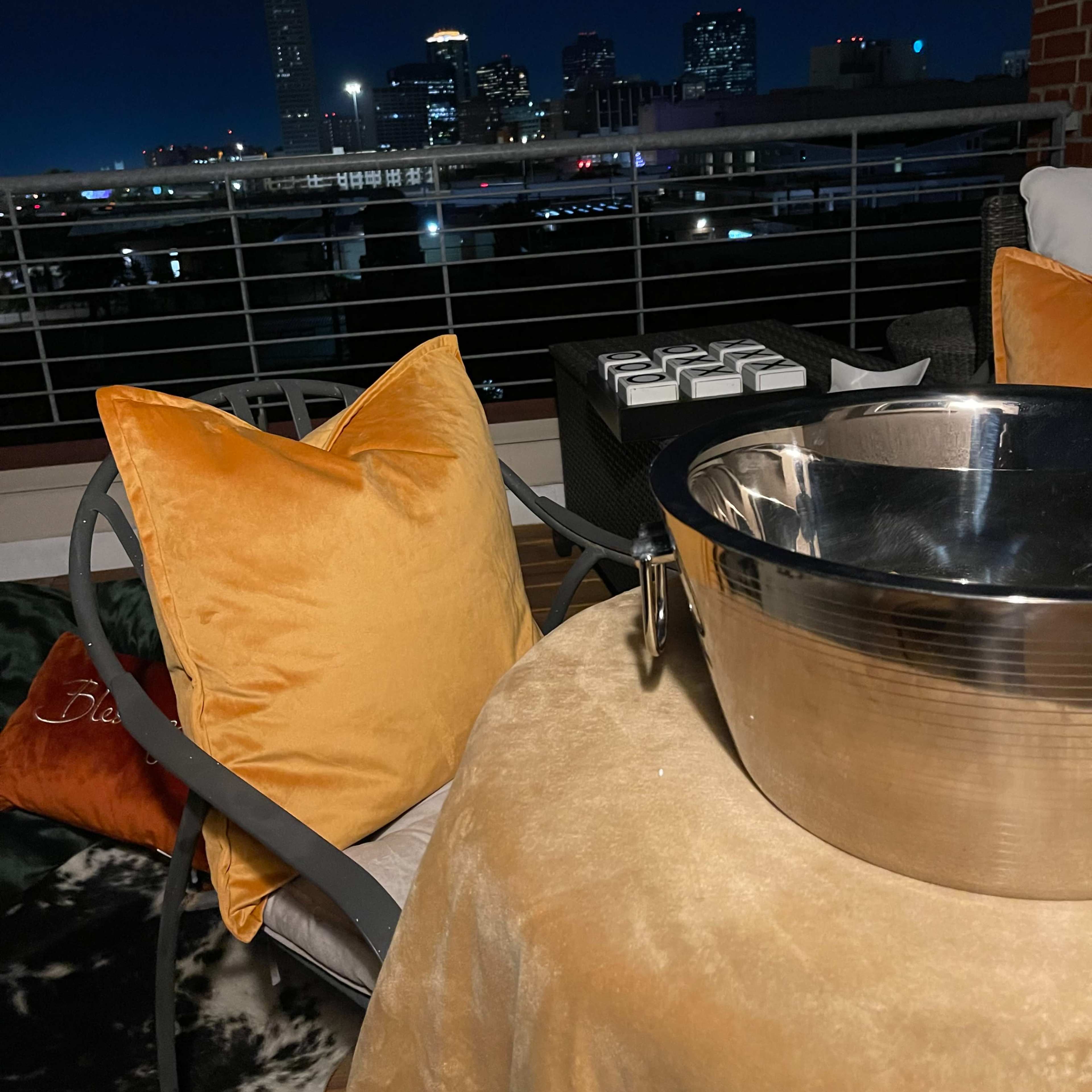 A balcony setting with a metal bowl and orange cushions overlooking a city skyline at night.