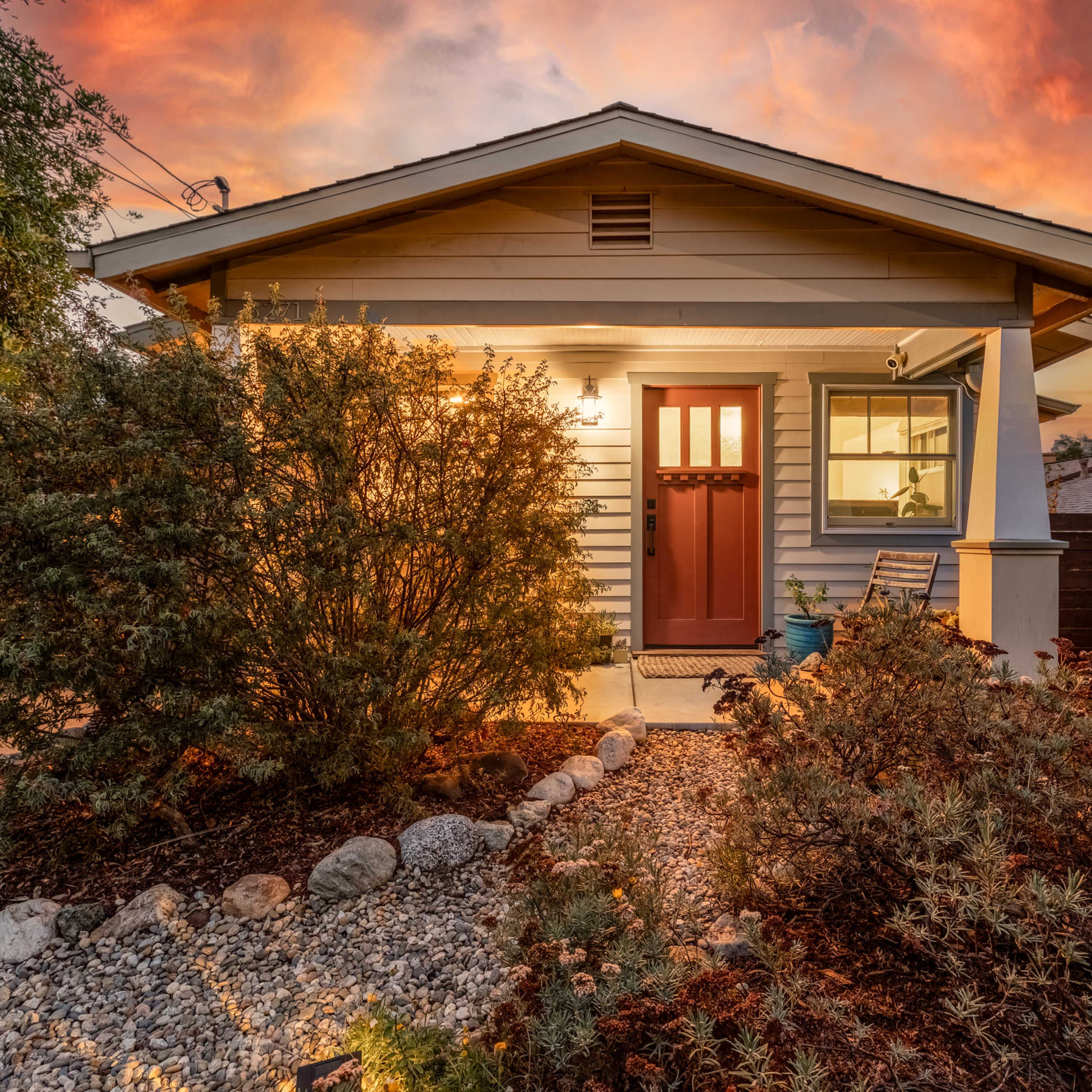 A small house with a brown front door is surrounded by landscaped gardens and rocks under a colorful sunset sky.