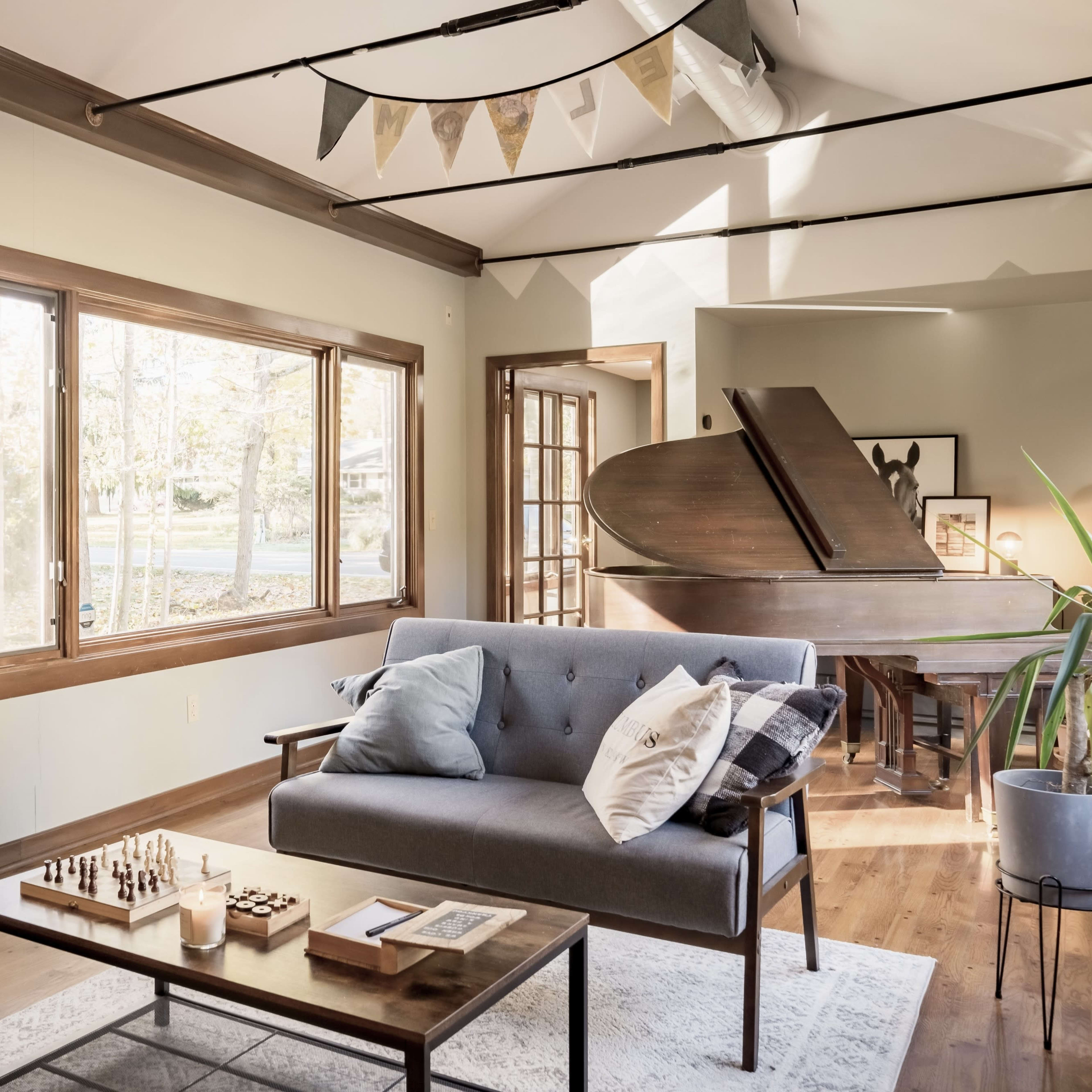 A cozy living room features a gray sofa, a wooden coffee table with a chess set, and a grand piano near large windows that let in natural light.