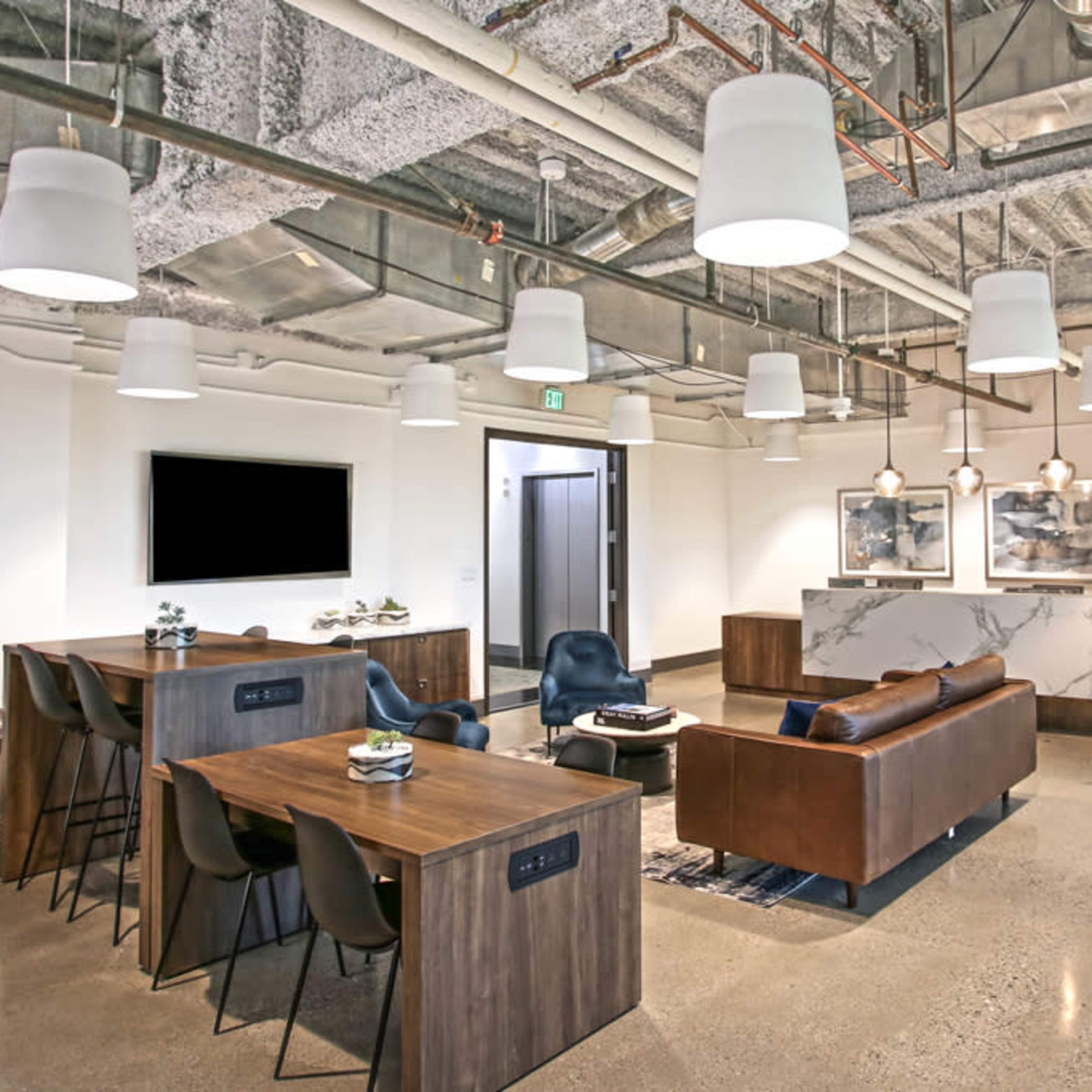 A modern office lounge area featuring a seating arrangement with a sofa and chairs, several tables, and exposed ceiling ducts.