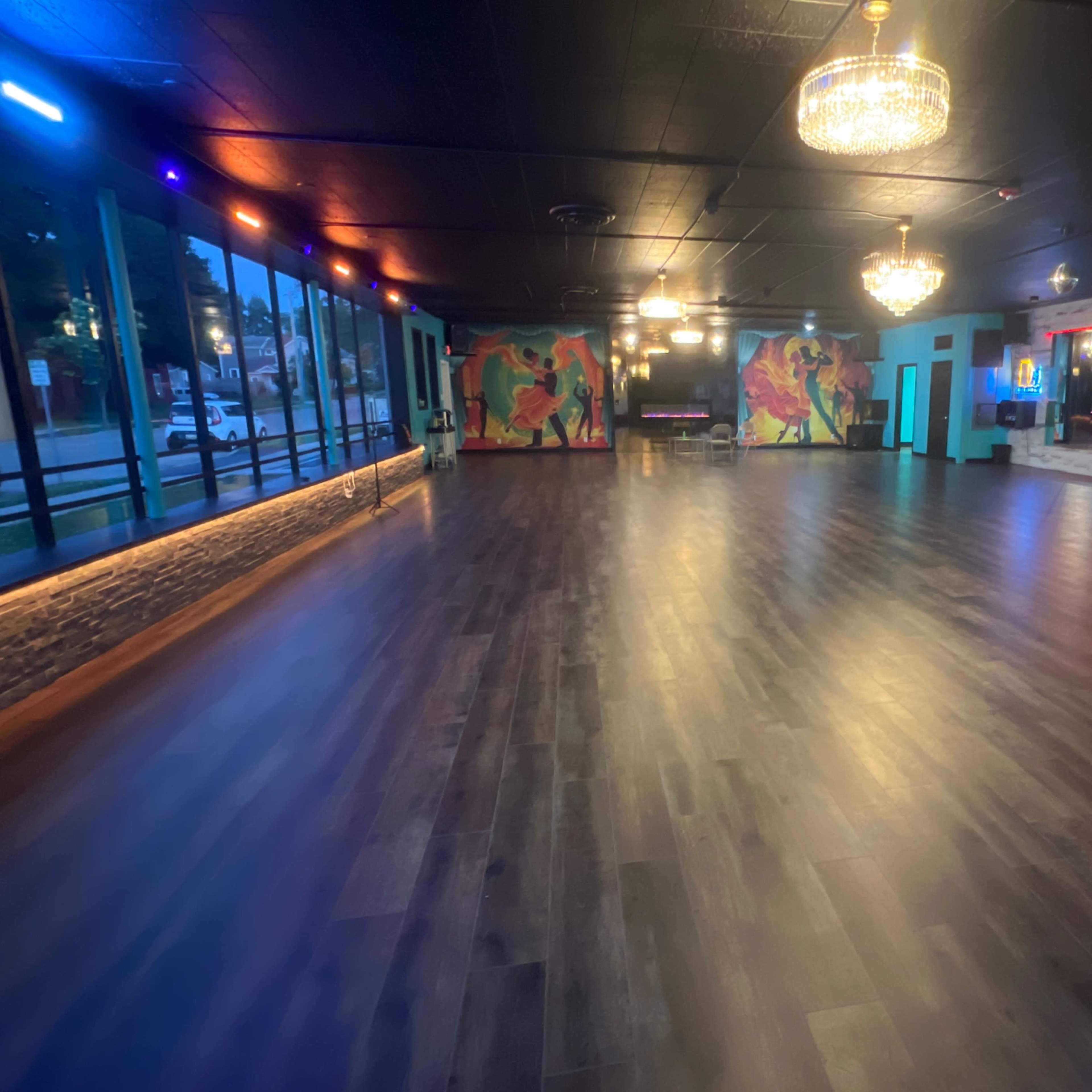 A spacious dance studio with wooden floors, colorful murals on the walls, and dim lighting accented by chandeliers.