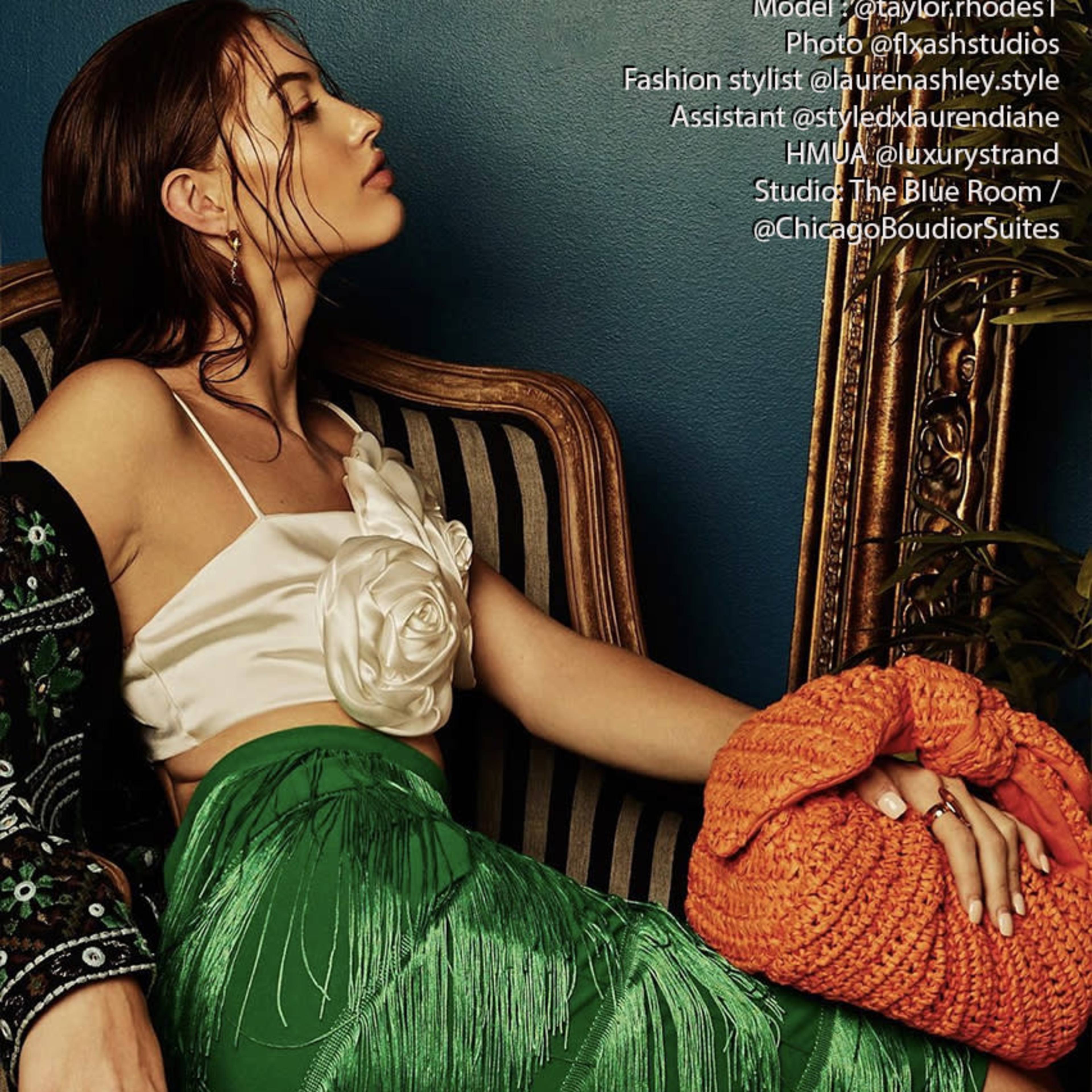 A model in a white top adorned with a floral embellishment sits on a striped chair, wearing a green fringed skirt and holding an orange bag.