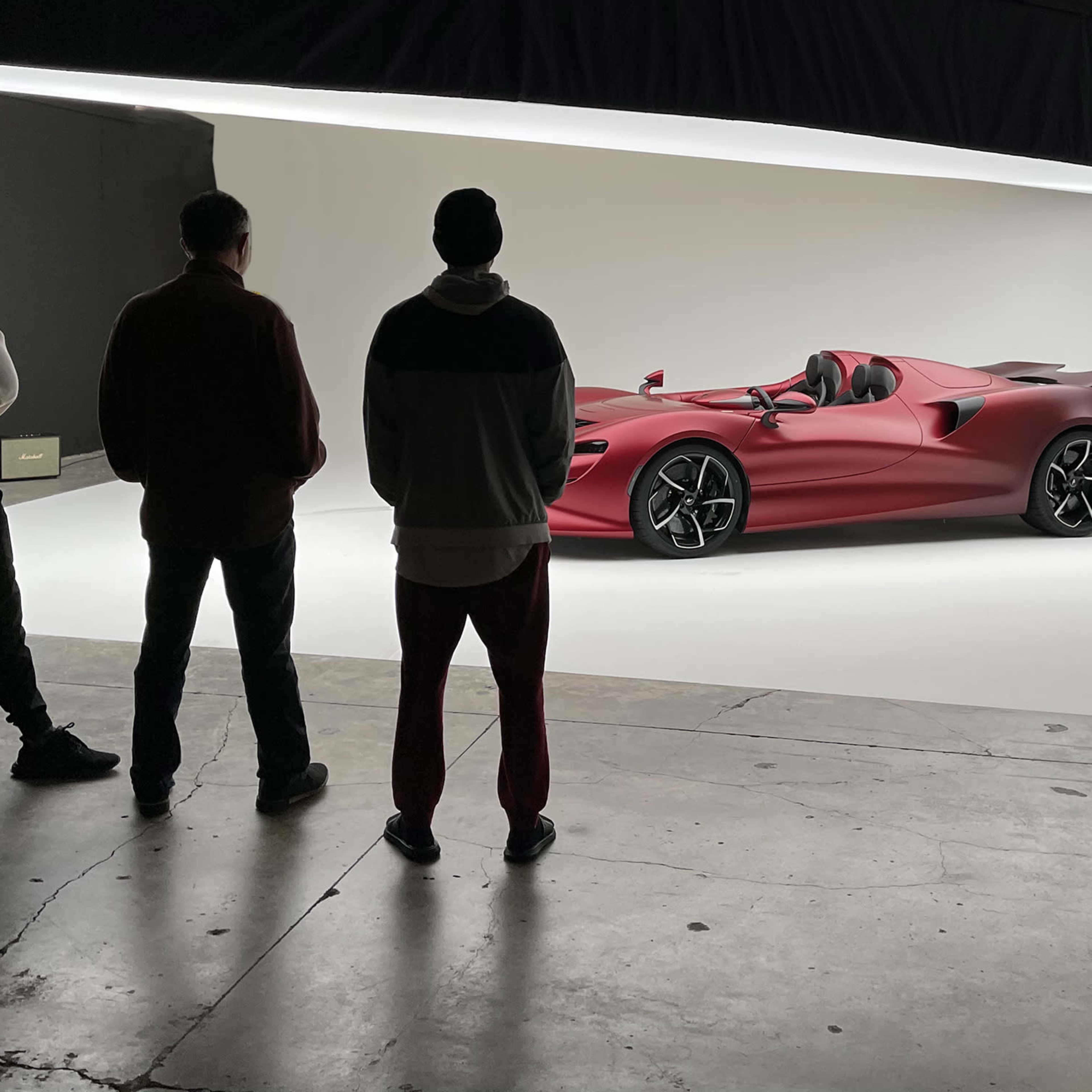 A group of five people stands in a studio, observing a sleek red sports car positioned on a white backdrop.