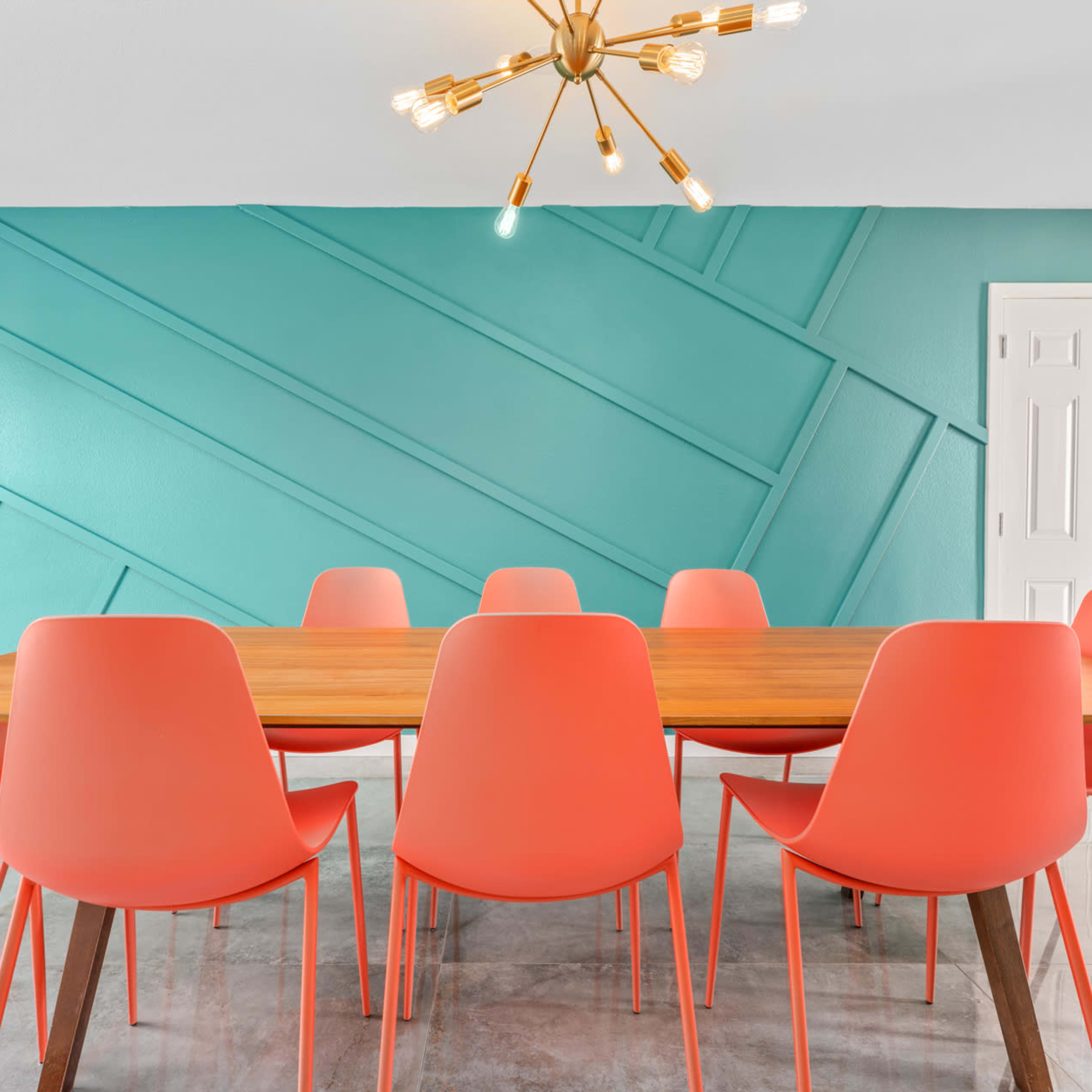A modern dining room features a large wooden table surrounded by eight coral-colored chairs against a turquoise accent wall.