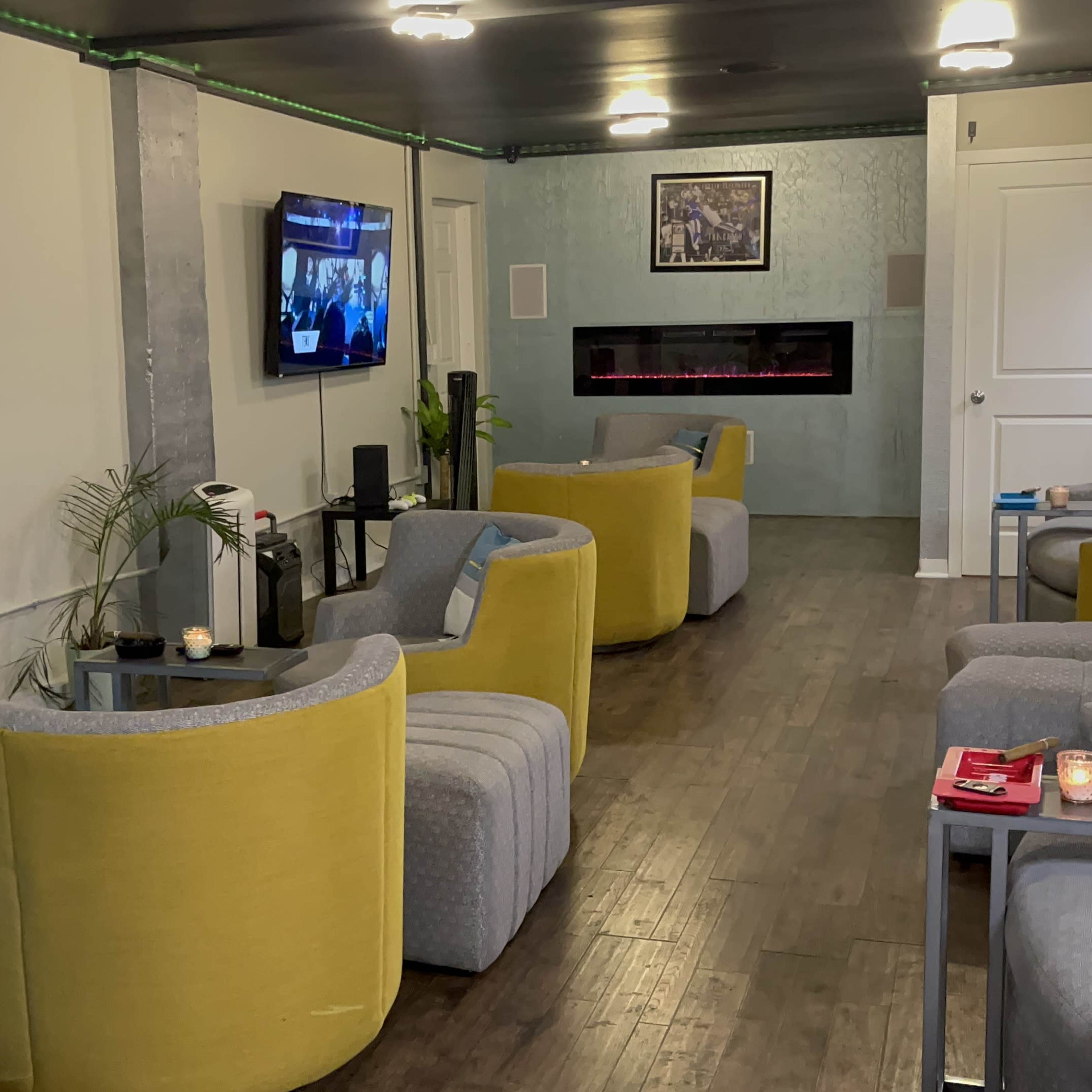 A modern waiting area with yellow and gray armchairs, two televisions, a faux fireplace, and a plant.