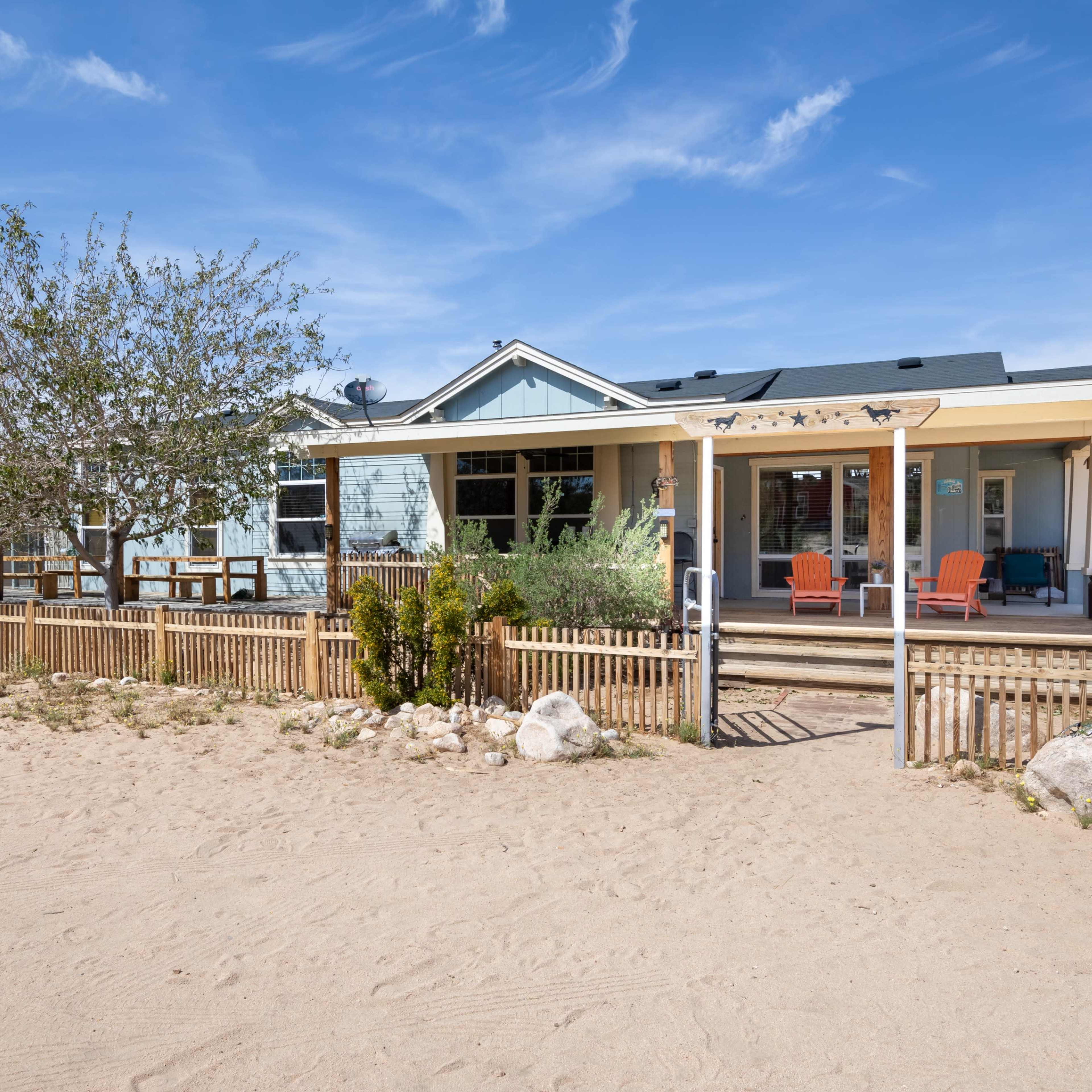 A single-story house with a porch and two orange chairs is surrounded by a wooden fence and desert landscaping.