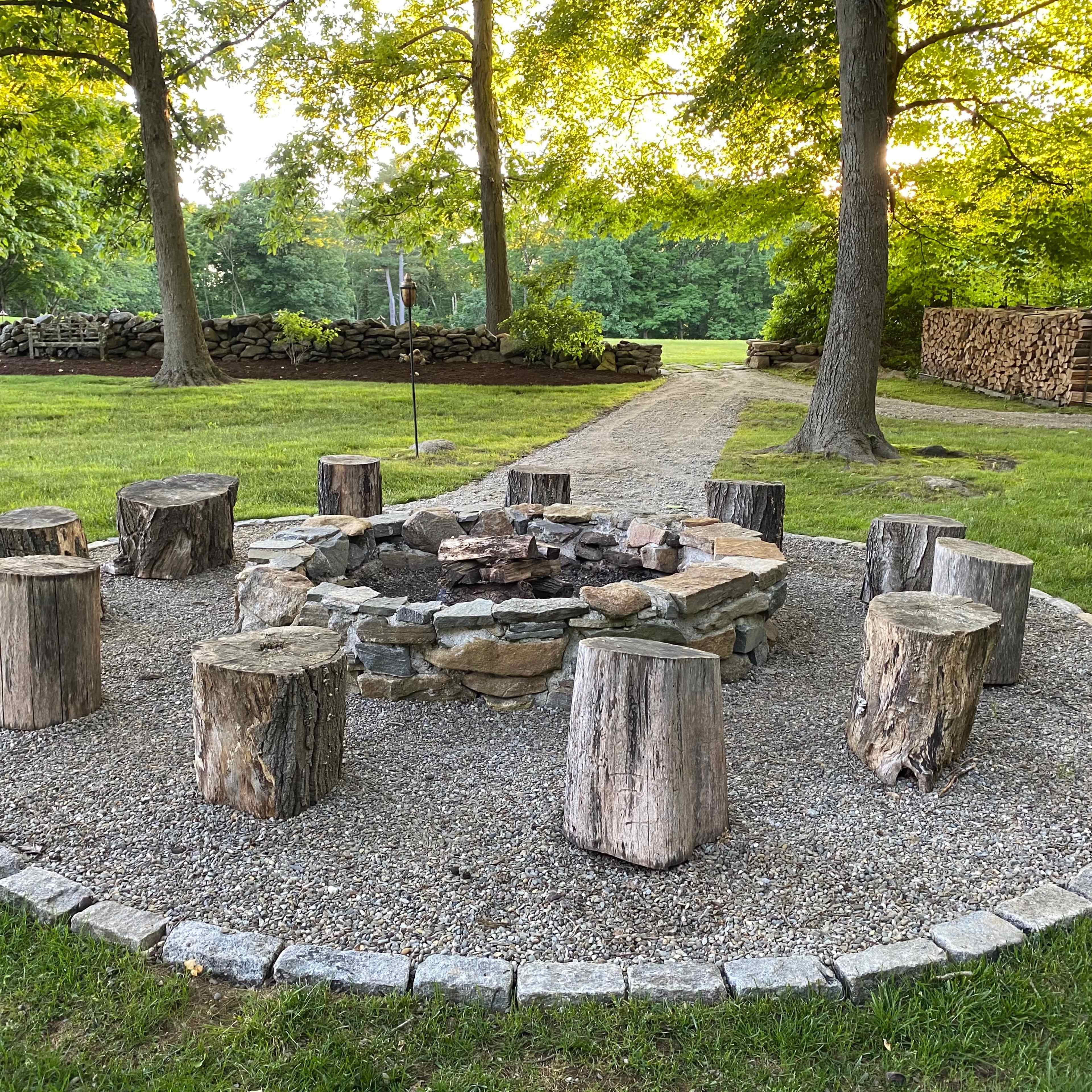 A circular fire pit made of stones surrounded by wooden logs arranged as seating, set within a grassy area with trees and stacked firewood in the background.