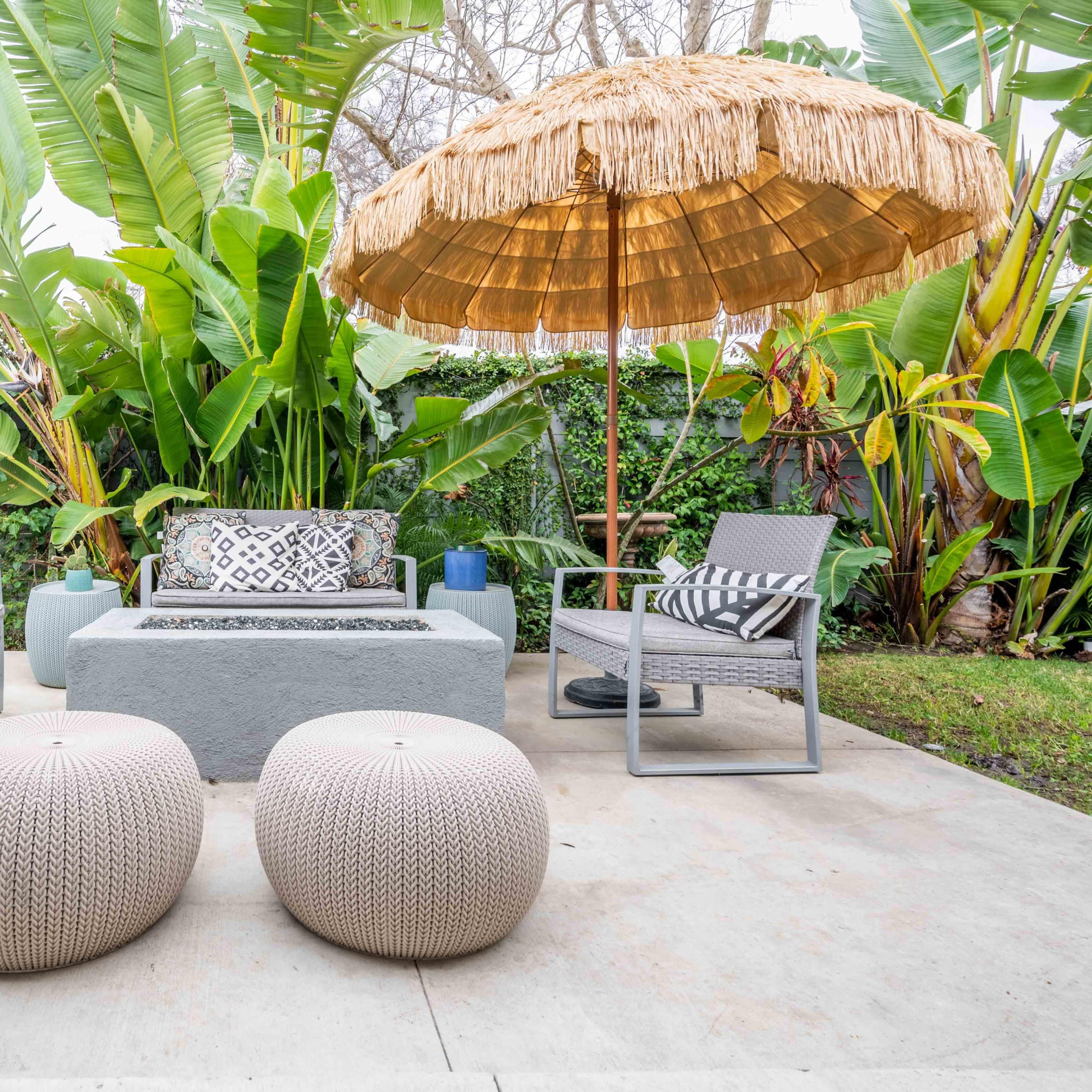 A patio area features a gray stone table surrounded by two chairs and two round ottomans, all under a thatched umbrella surrounded by lush greenery.