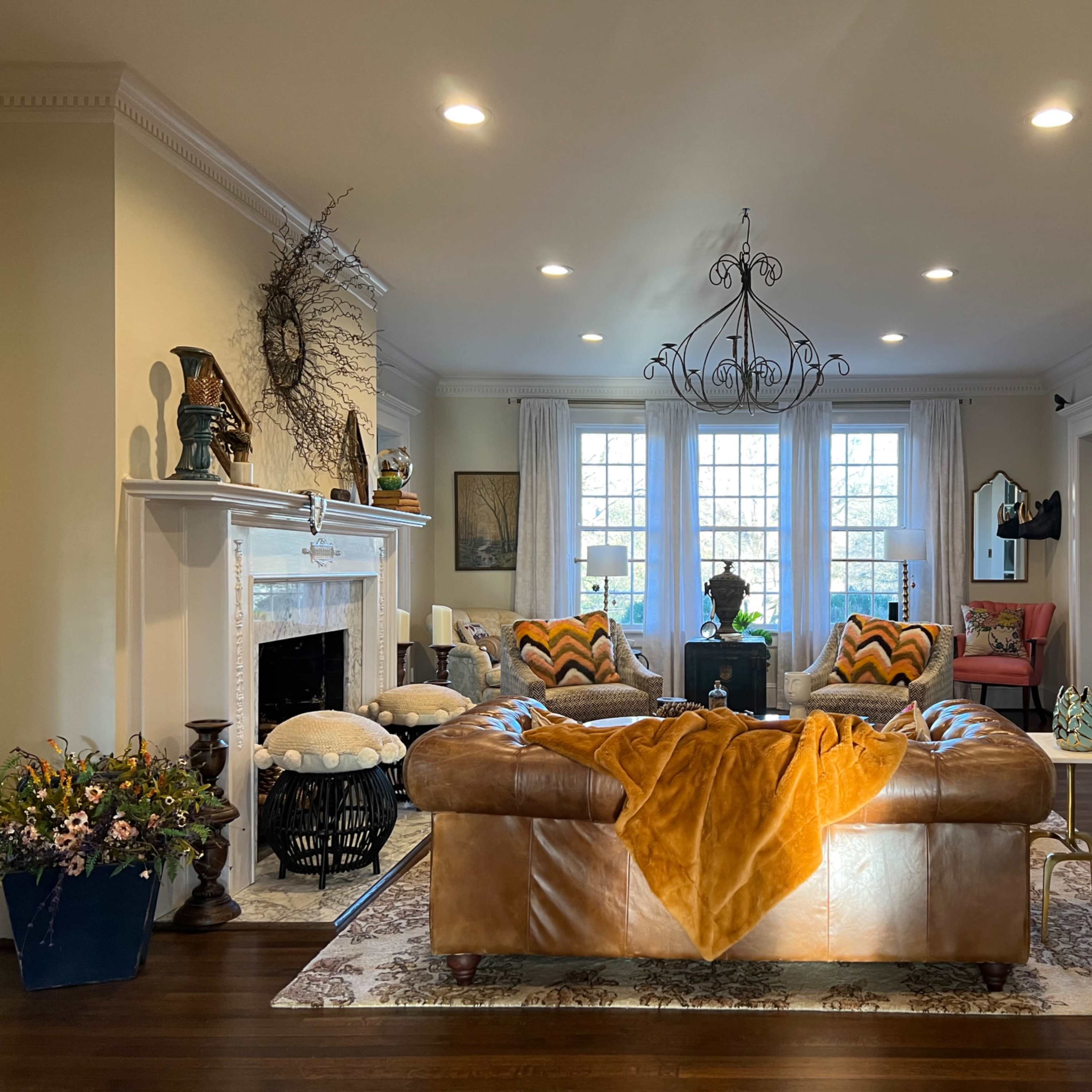 A spacious living room features a brown leather sofa, a decorative chandelier, and large windows letting in natural light, surrounded by various plants and artwork.