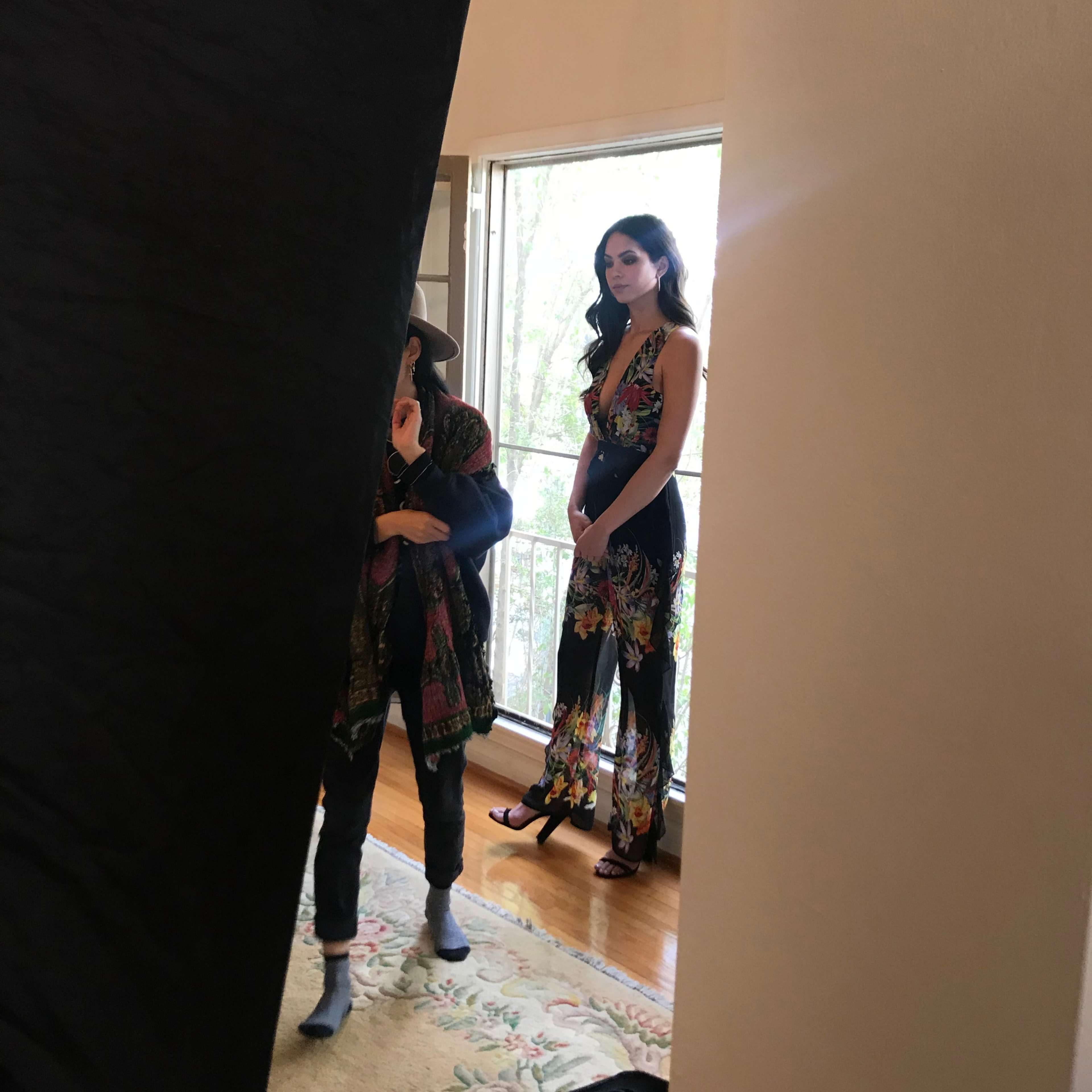 A model wearing a floral jumpsuit stands near a window while a photographer adjusts equipment in a well-lit room.
