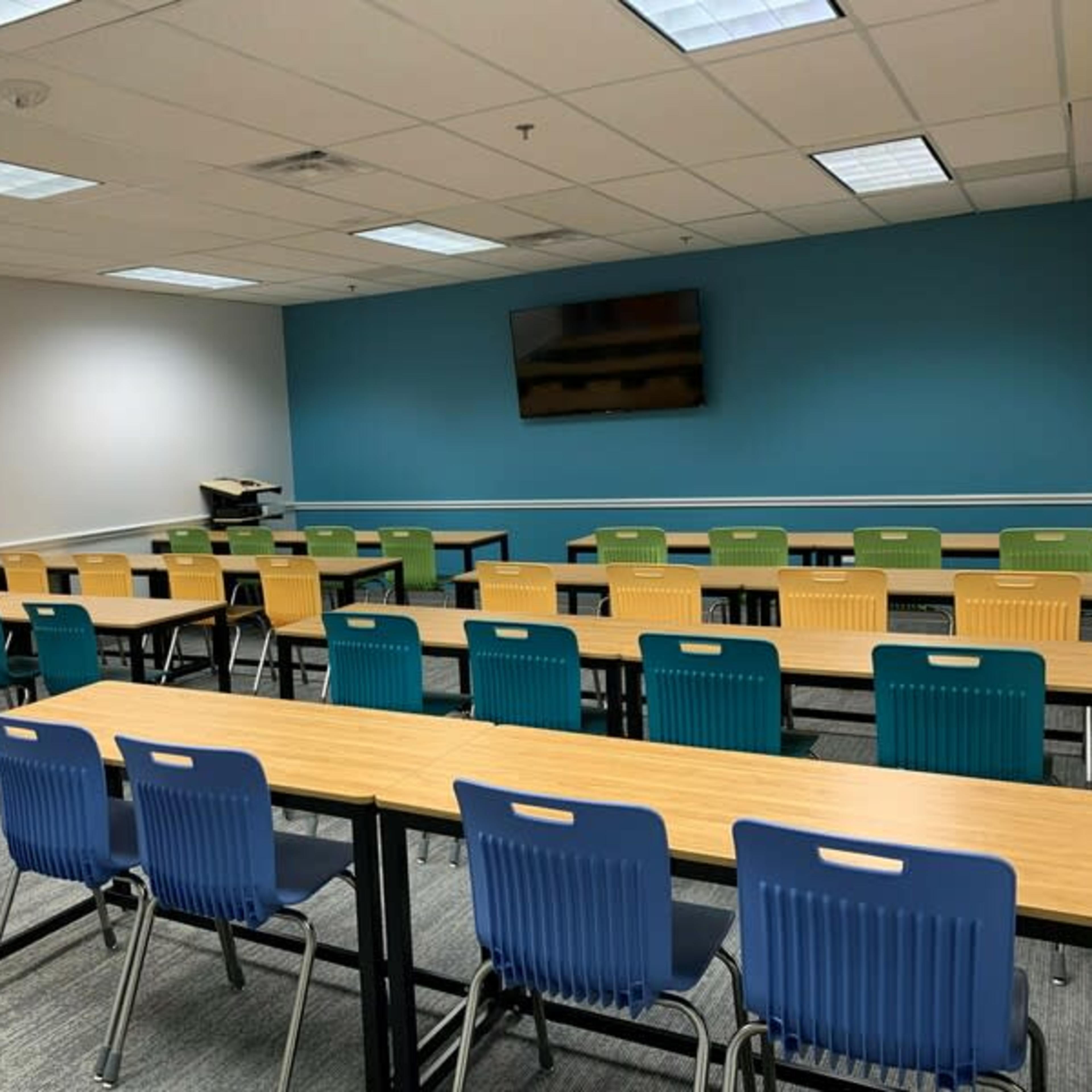 A classroom features several rows of tables with colorful chairs and a wall-mounted television.