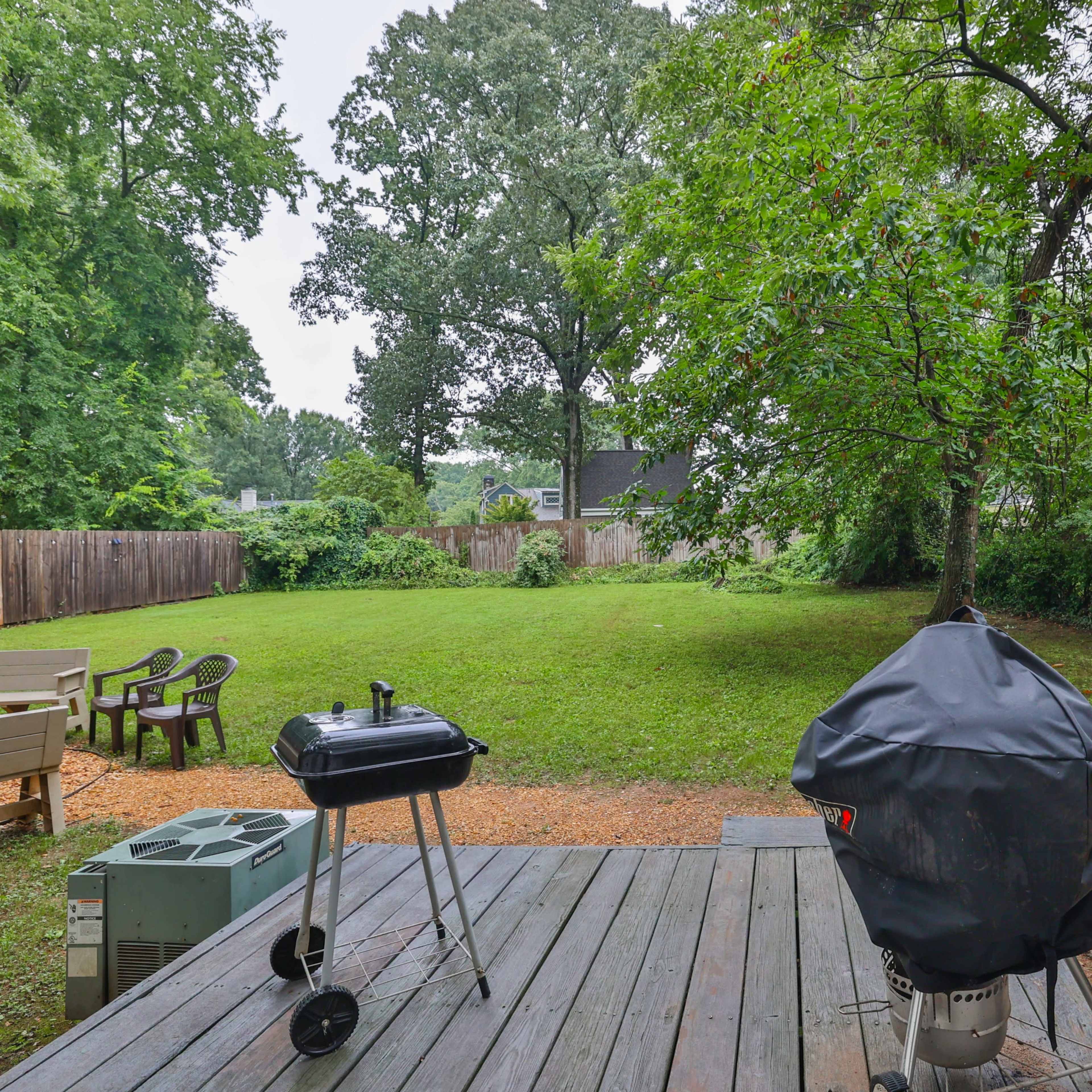 A backyard with a wooden deck, a gas grill, and a charcoal grill, surrounded by green grass and trees.