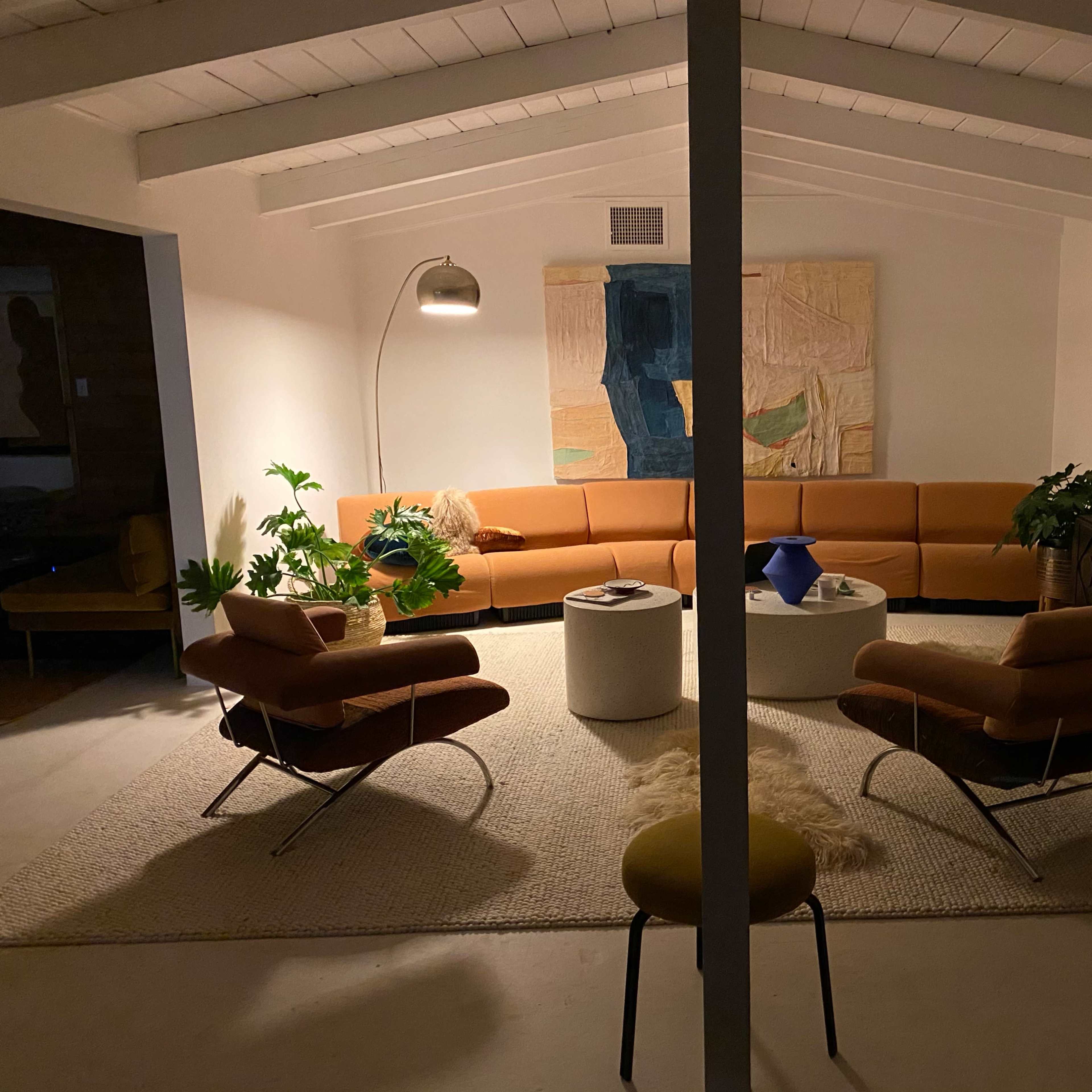 A modern living room with a curved orange sofa, two brown armchairs, a round white coffee table, and various plants, all under a ceiling with wooden beams.