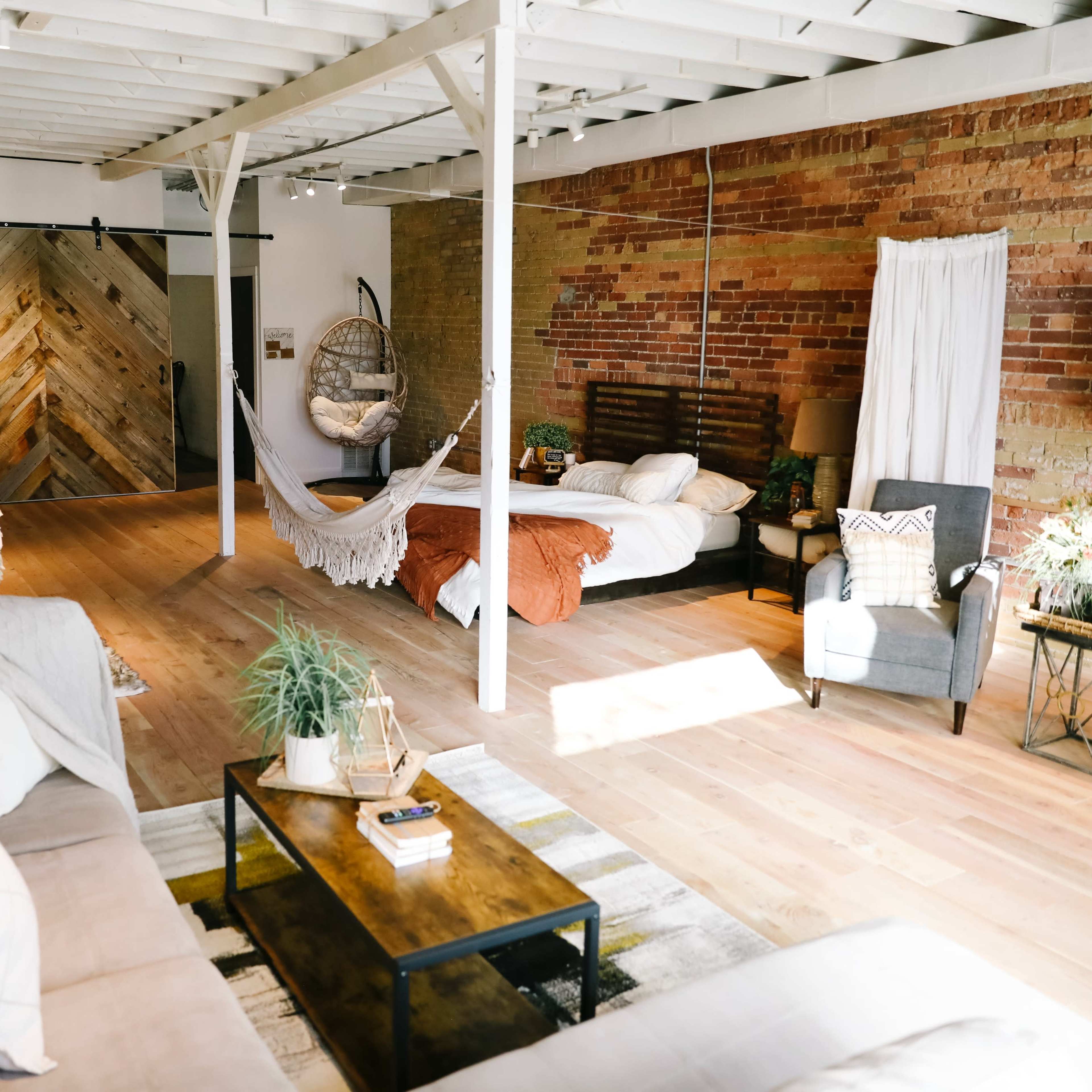 A spacious room featuring a mix of rustic brick and wooden walls, with a large bed, a sofa, and a hanging chair.