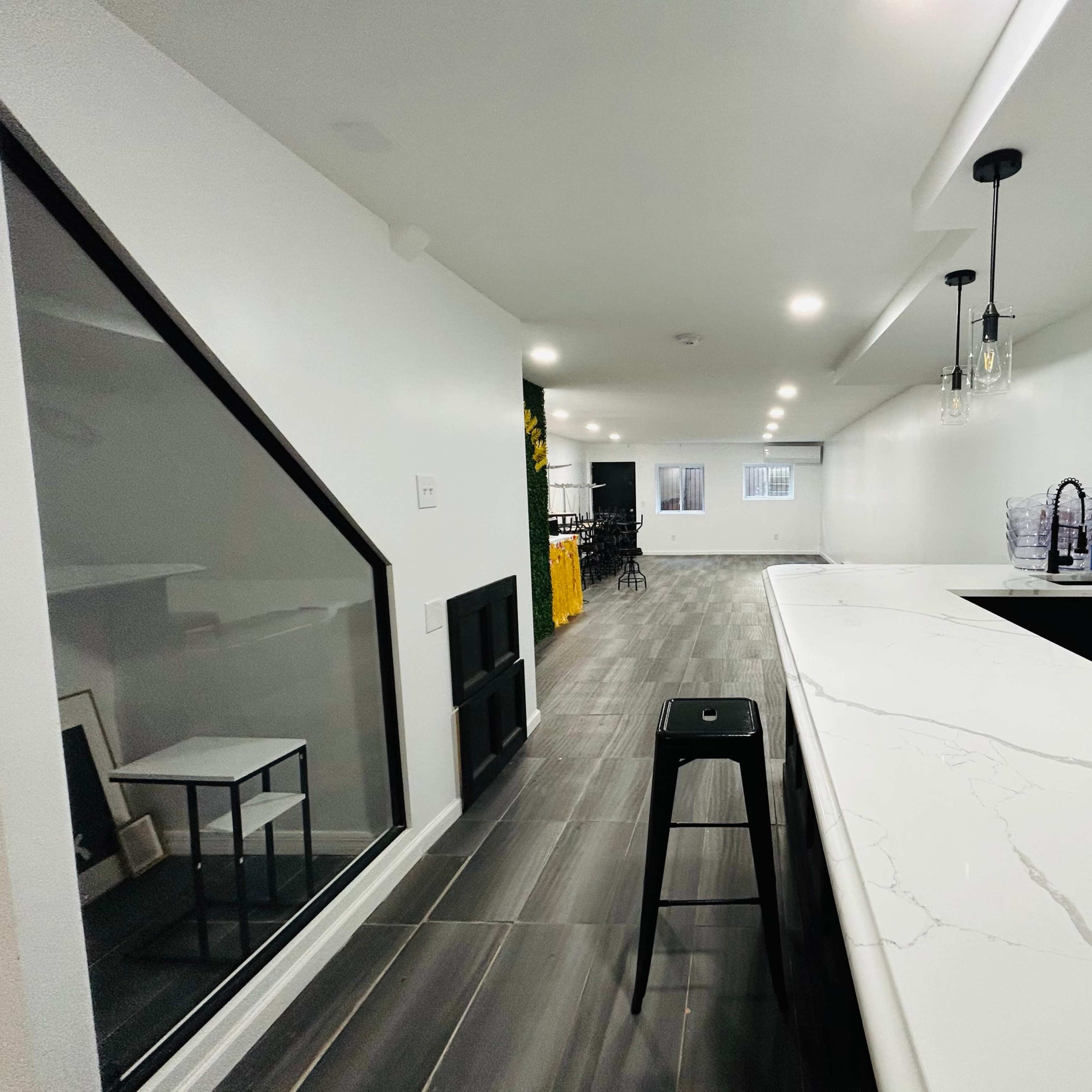 A modern, open-concept interior space with a long white countertop bar, black stool, and large tiled flooring, leading to a seating area in the background.