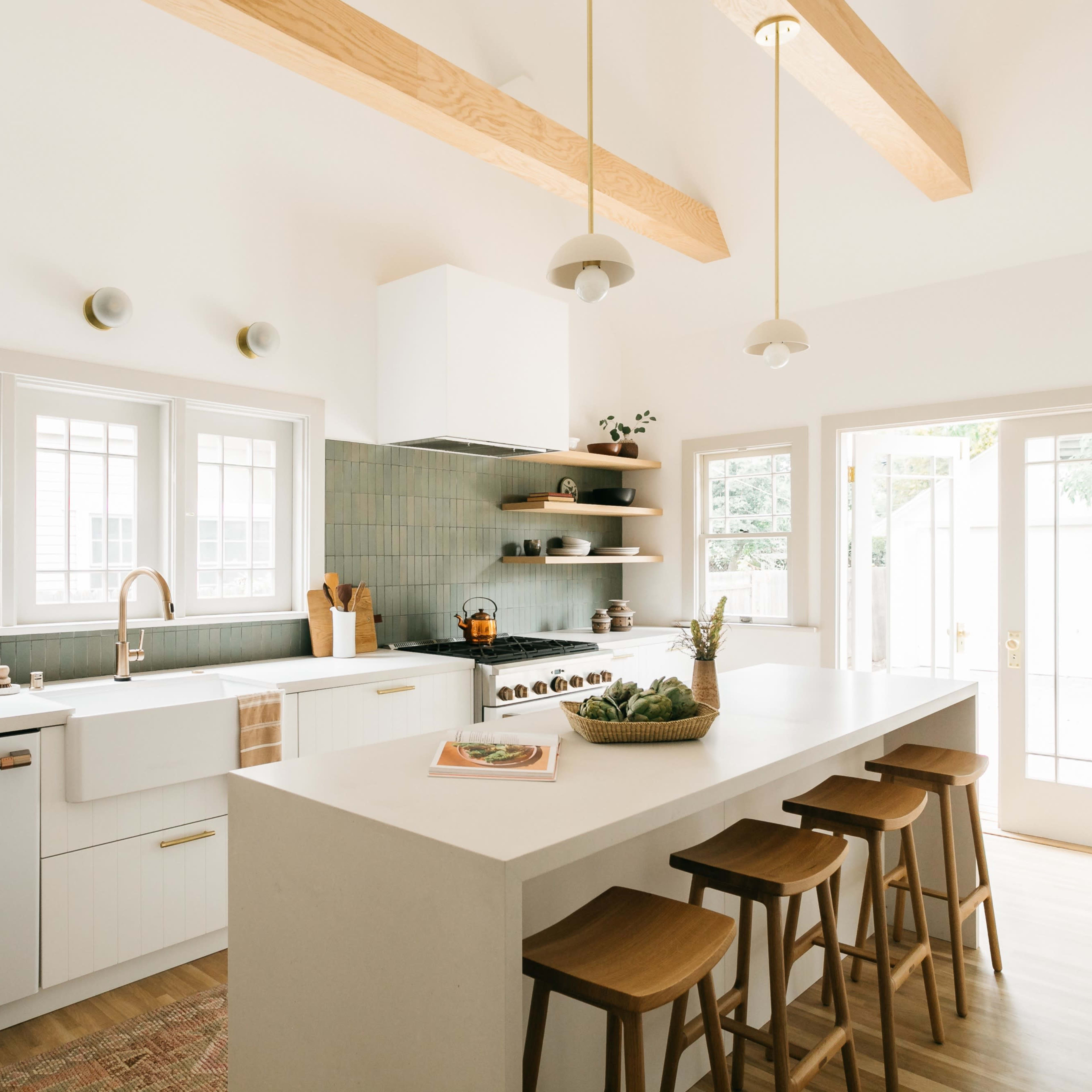A modern kitchen features a large island with seating, light wood beams on the ceiling, and green tile backsplash behind a white sink and stove.