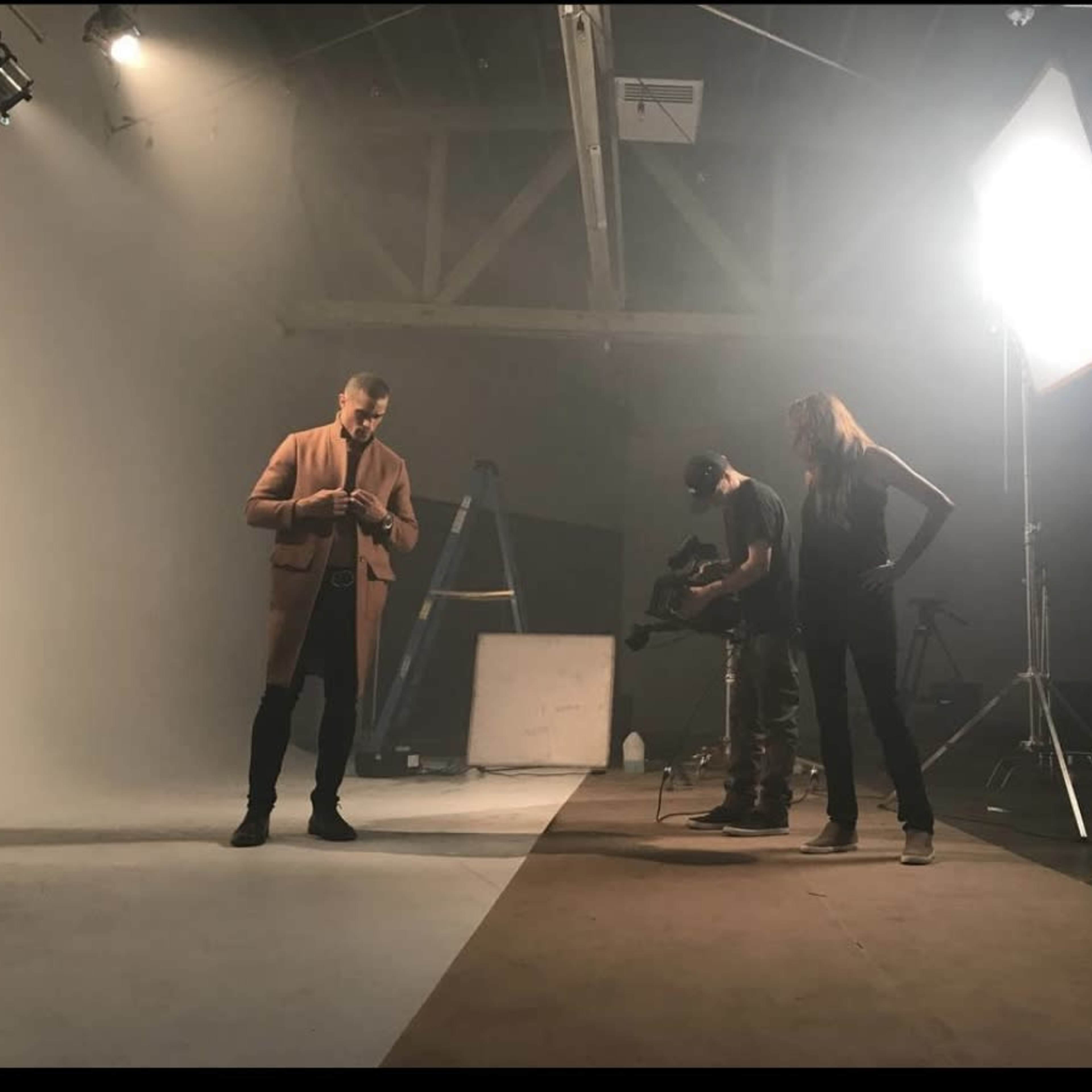A man in a long coat stands on a set while three crew members operate equipment in a well-lit studio.