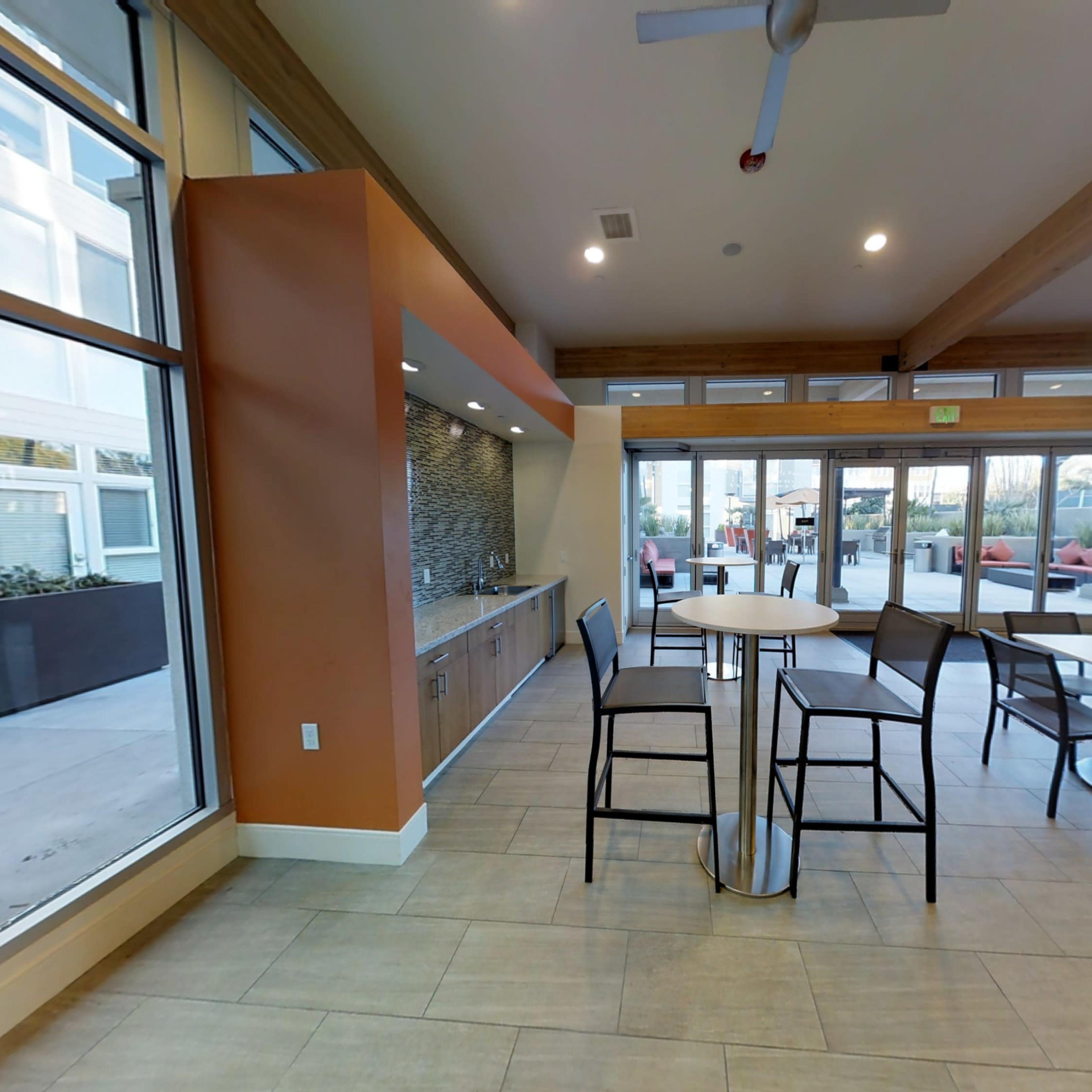 A modern indoor communal area with tall tables and chairs, large windows, and a kitchen space along one wall.
