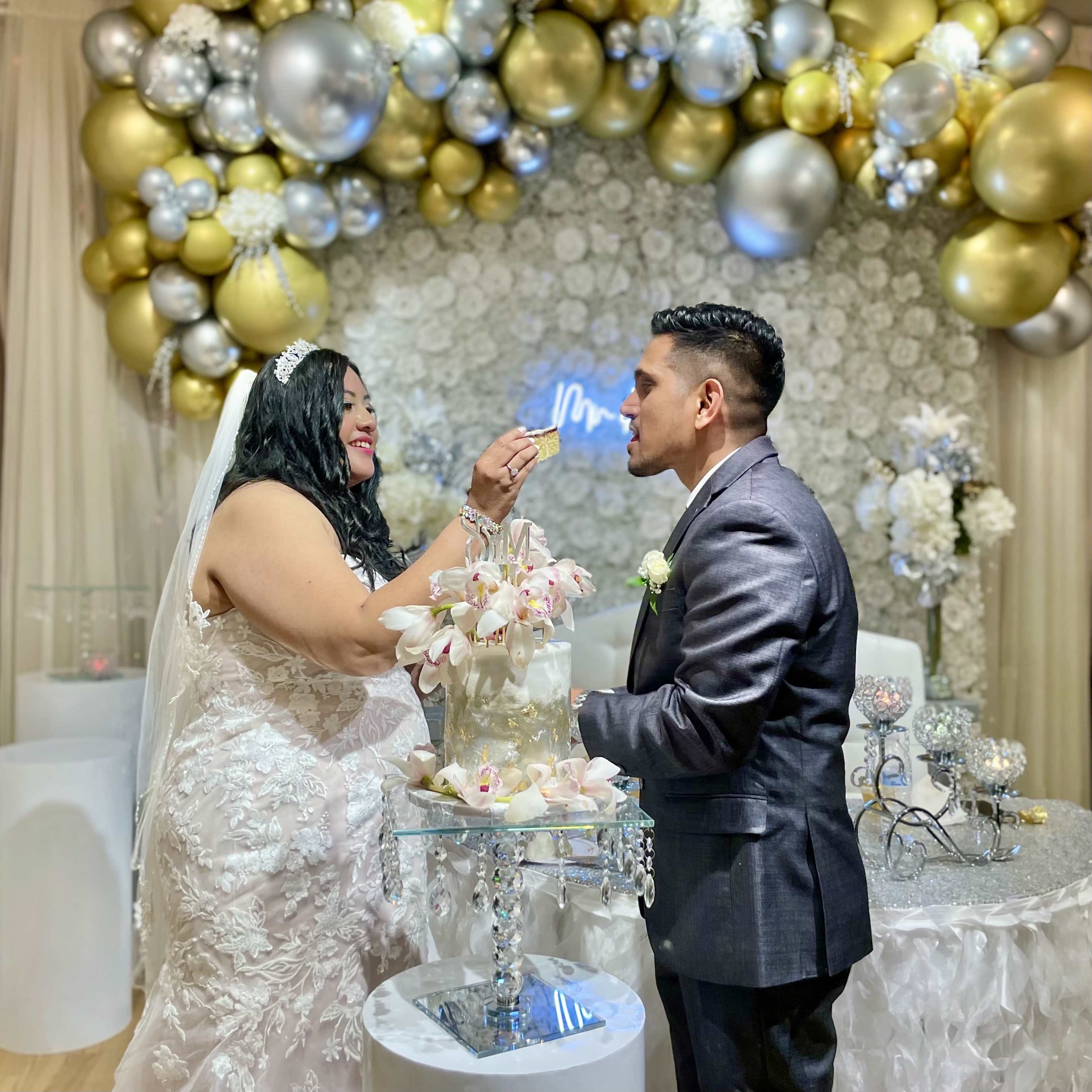 A bride and groom share a piece of cake in front of a decorated backdrop featuring balloons and floral arrangements.