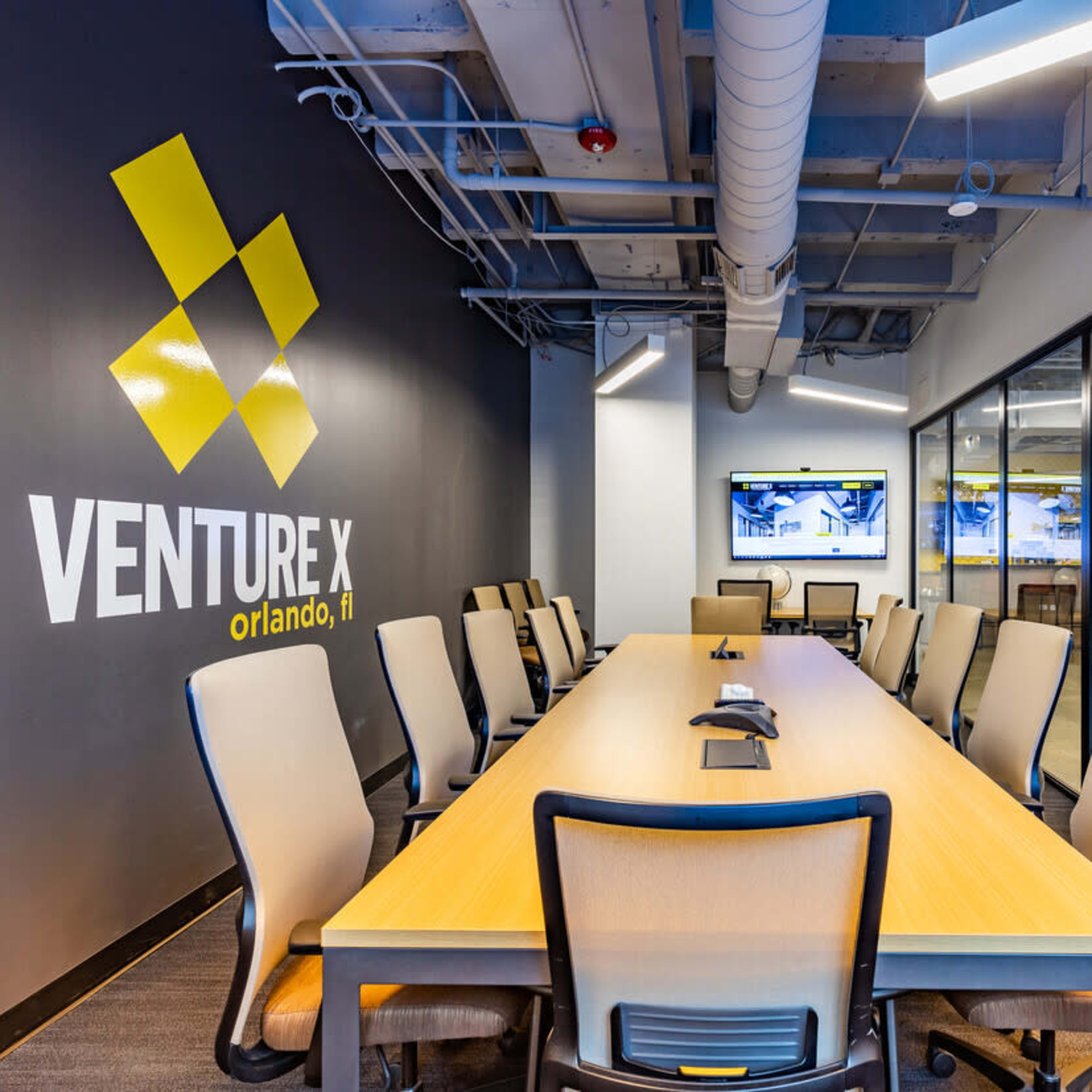 A modern conference room features a long wooden table surrounded by ergonomic chairs, with a large wall displaying the "Venture X" logo.