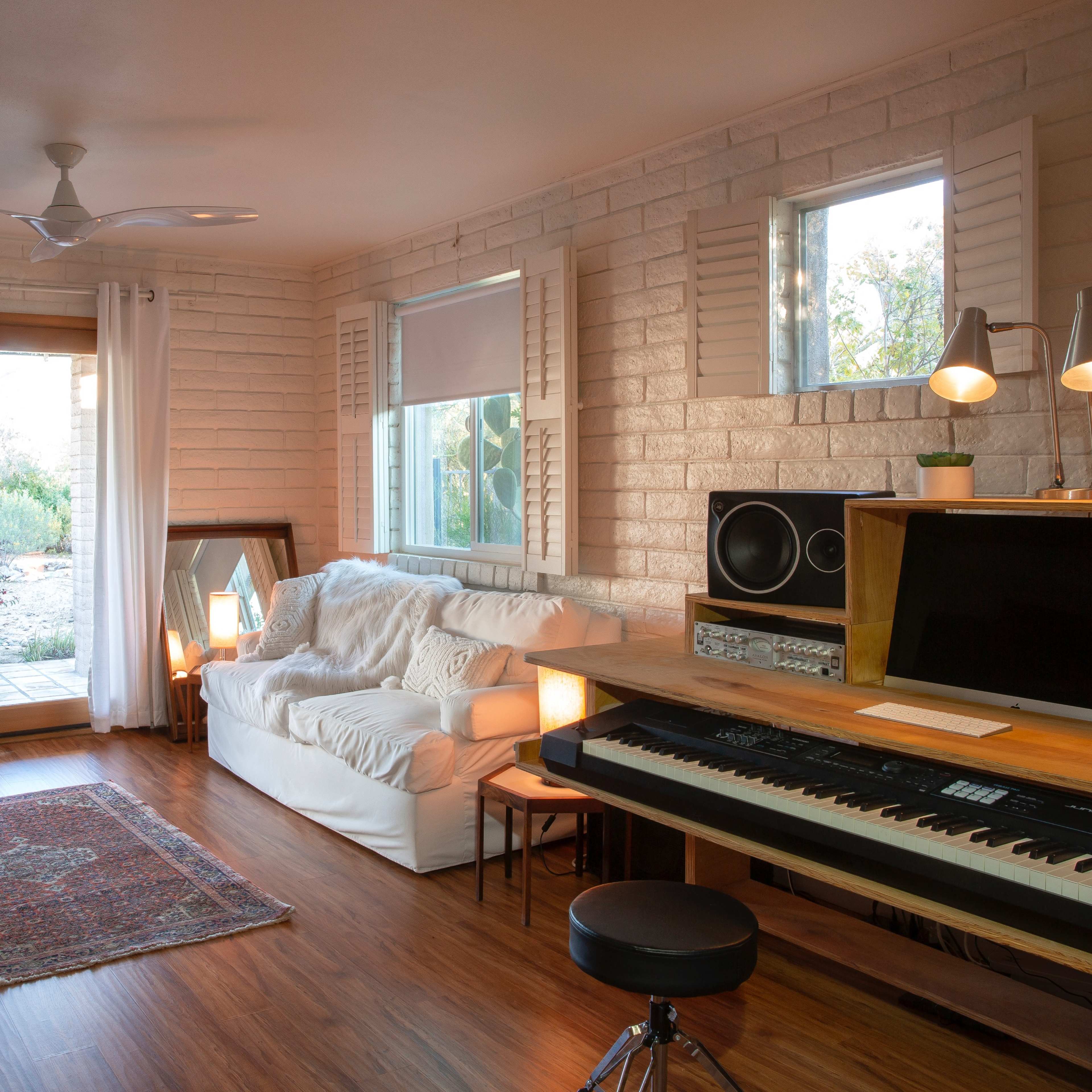 A bright living room features a white couch, a wooden desk with a piano, and large windows overlooking a patio.