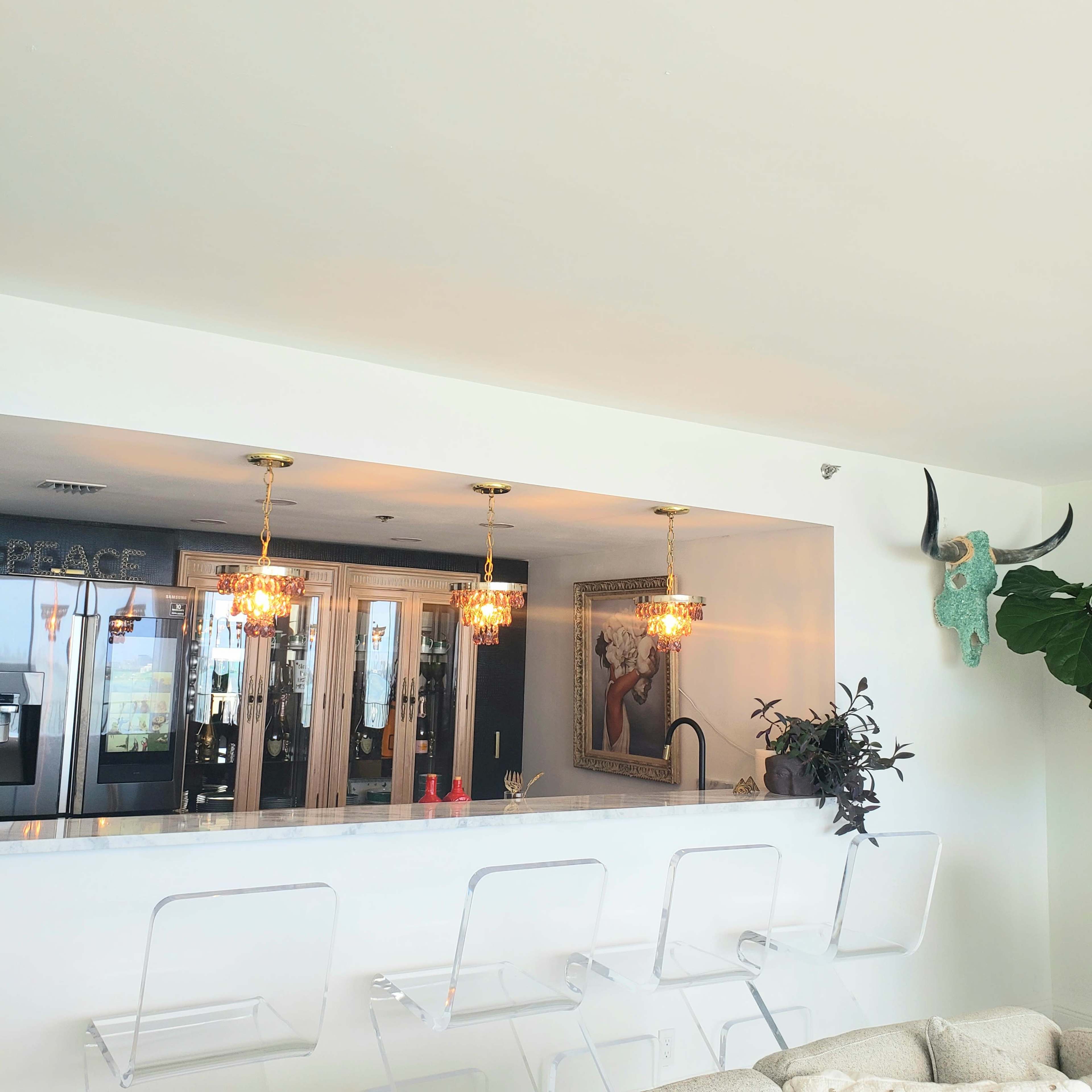 A modern kitchen bar with three transparent stools, illuminated by two ornate pendant lights, and featuring a decorative wall mounted antler and a potted plant.
