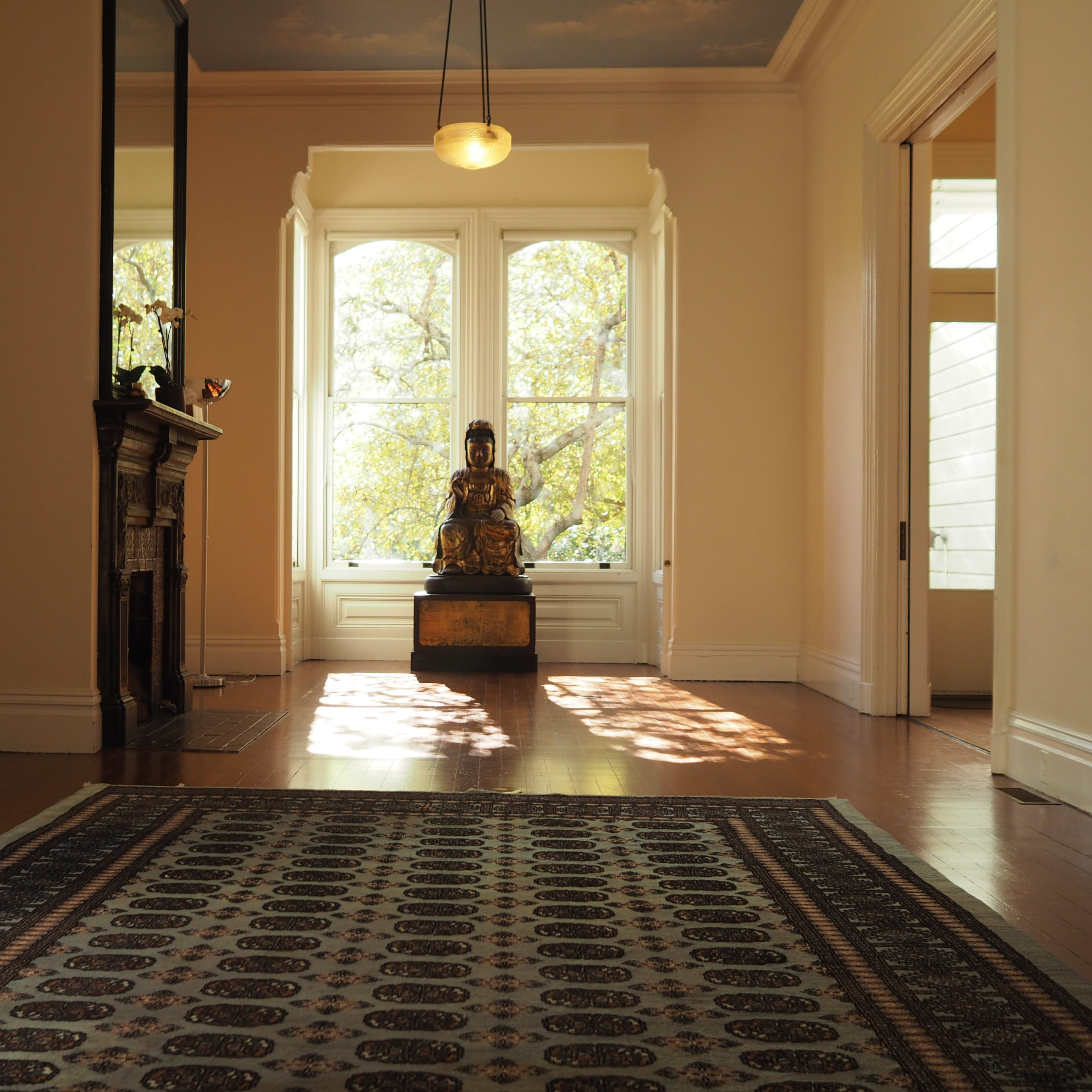 An empty room with a wooden floor, a decorative rug, large windows letting in natural light, and a seated Buddha statue positioned at the center.