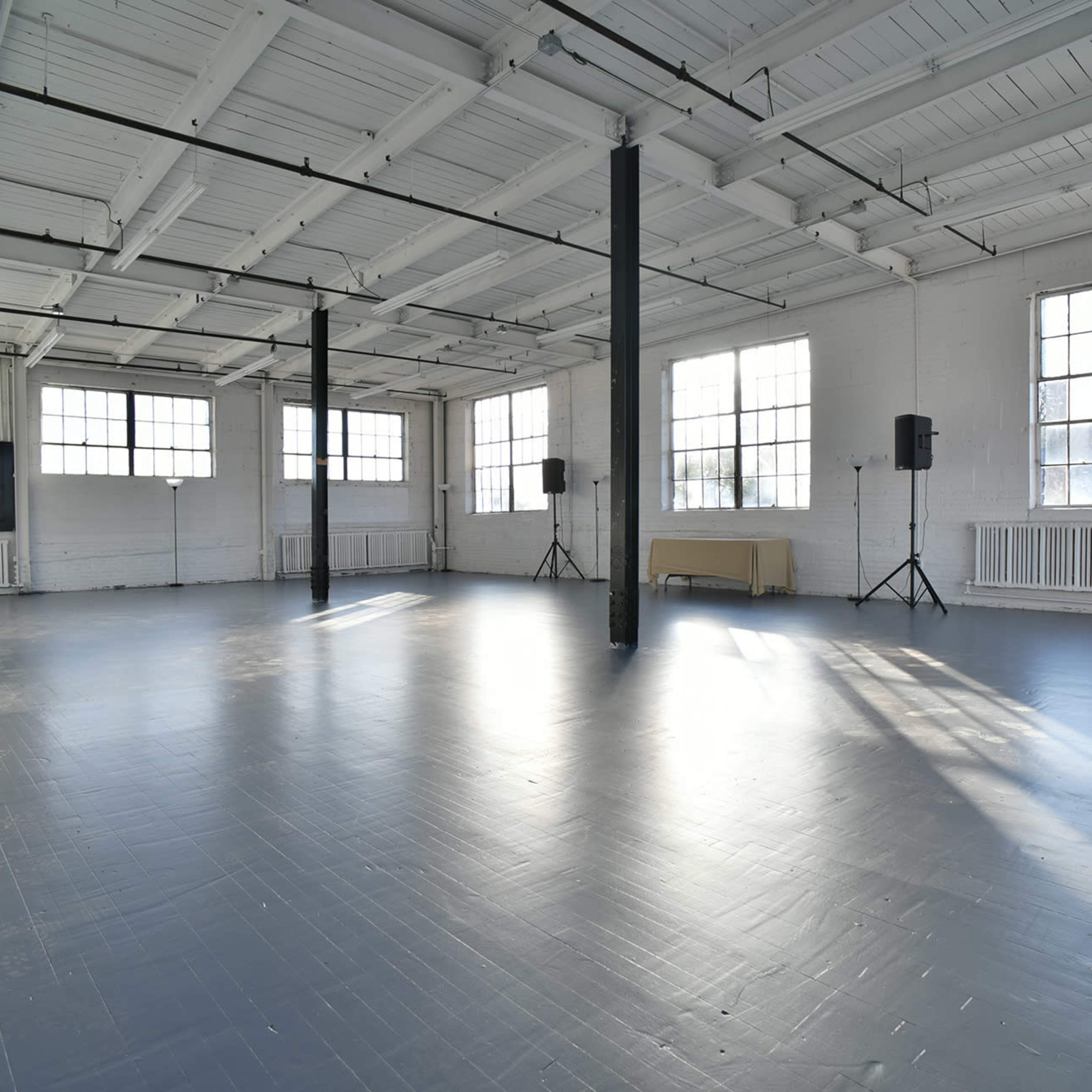 A spacious, well-lit interior with large windows and polished gray flooring, featuring two speakers on stands and a table against the wall.