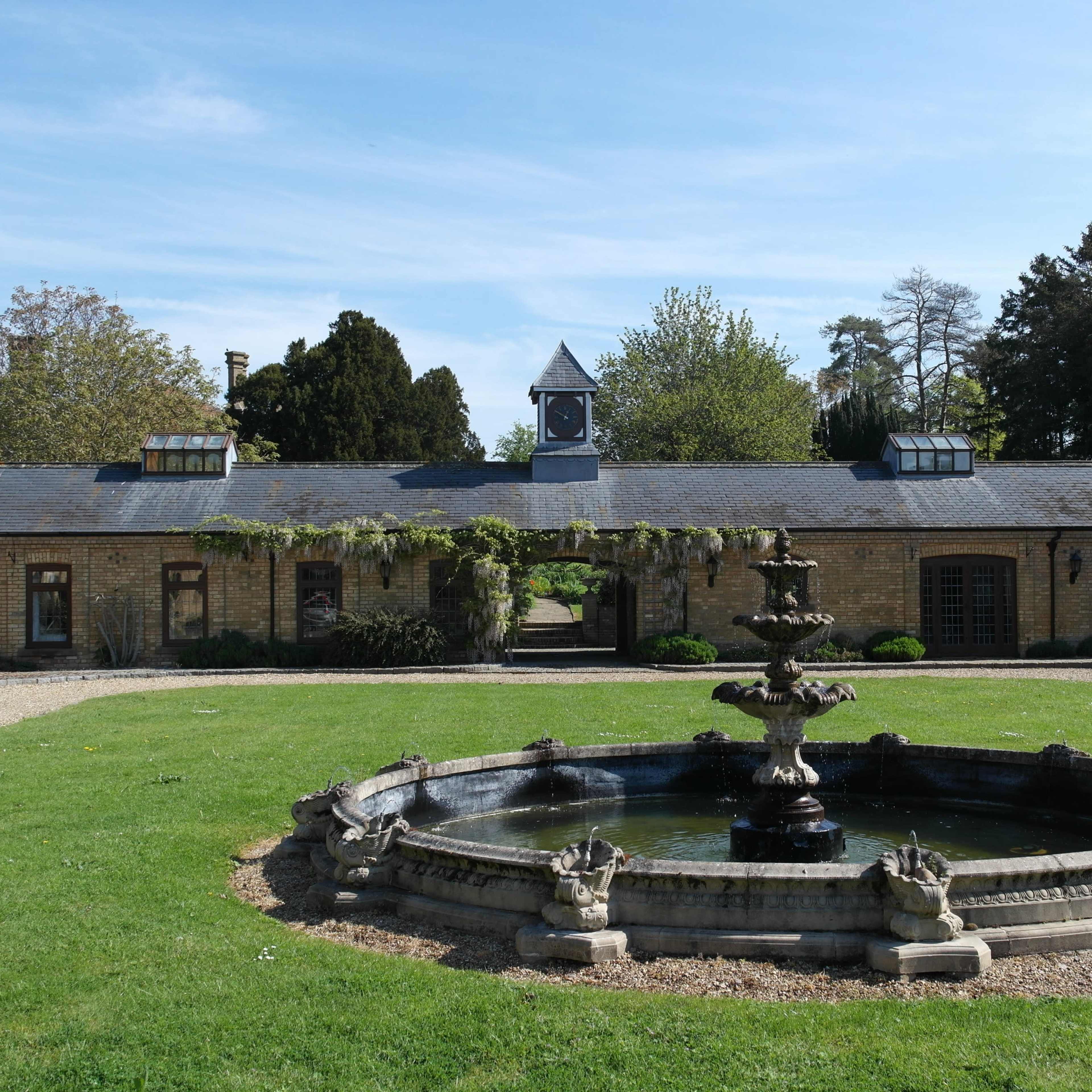 A large stone fountain sits in a grassy courtyard surrounded by buildings with windows and a clock tower in the background.