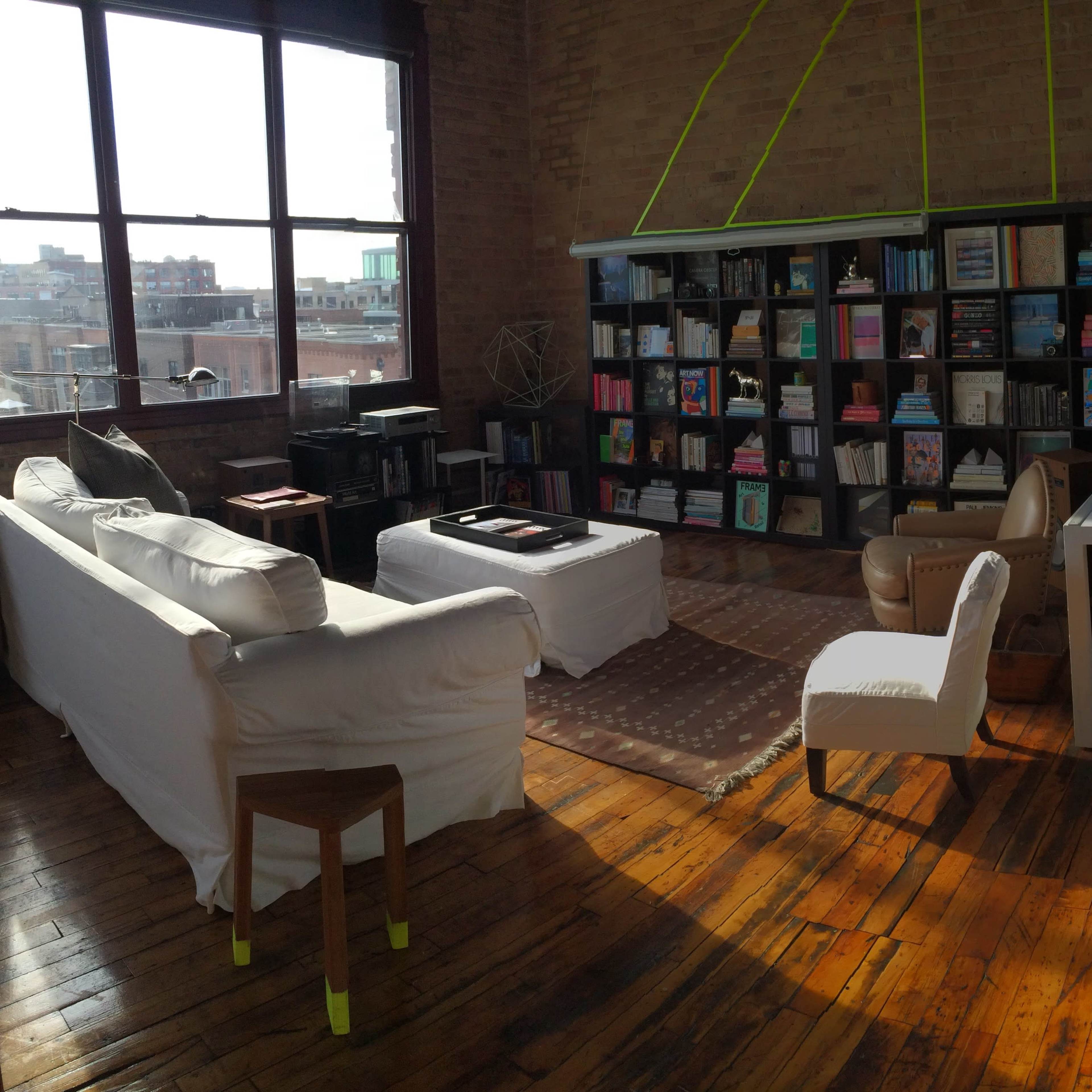 A spacious, sunlit living room with large windows, wooden floors, a white sofa, a bookshelf filled with books, and a simple desk and chair arrangement.