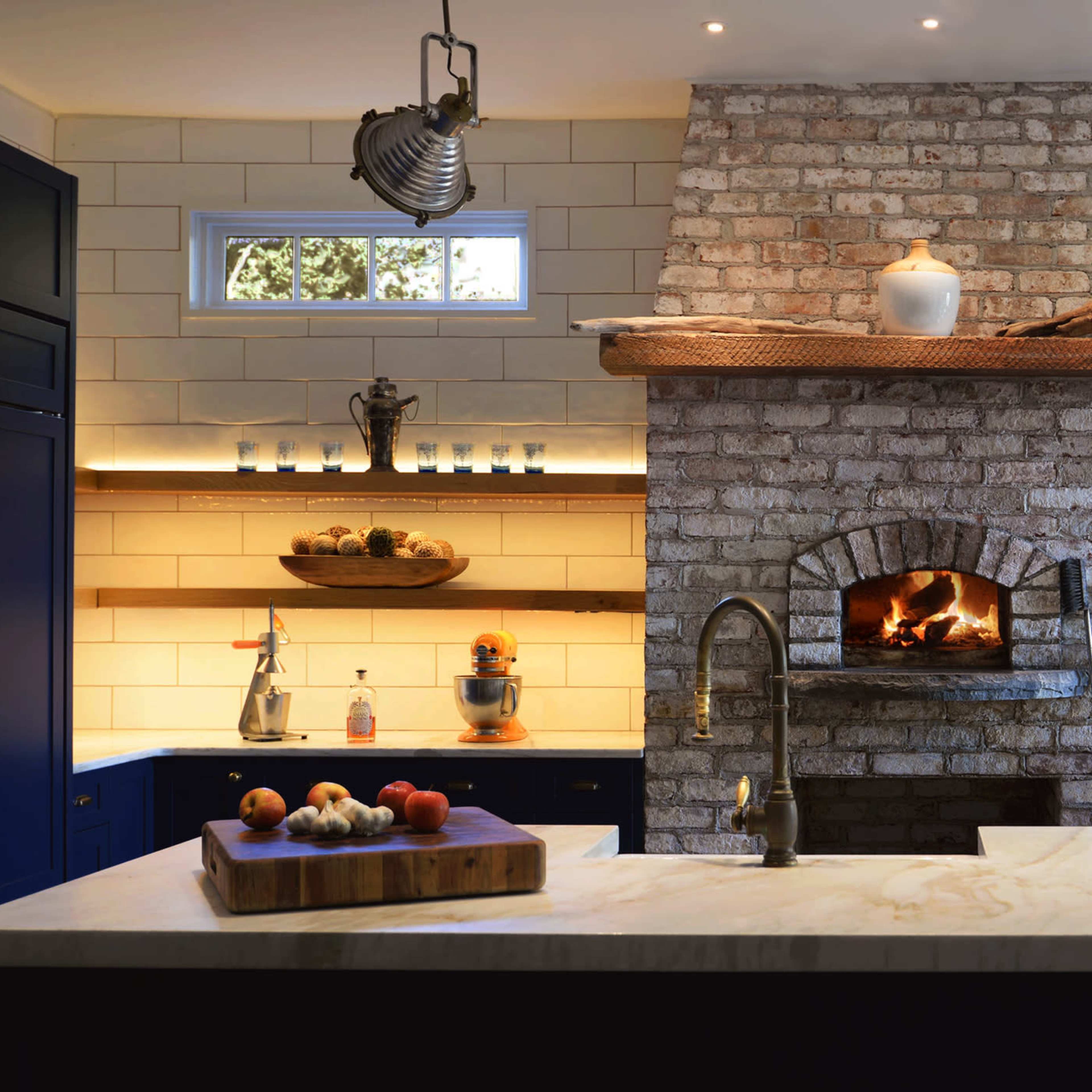 A modern kitchen features a large brick fireplace with a wooden mantel, stainless steel appliances, and open shelves displaying various kitchen items.