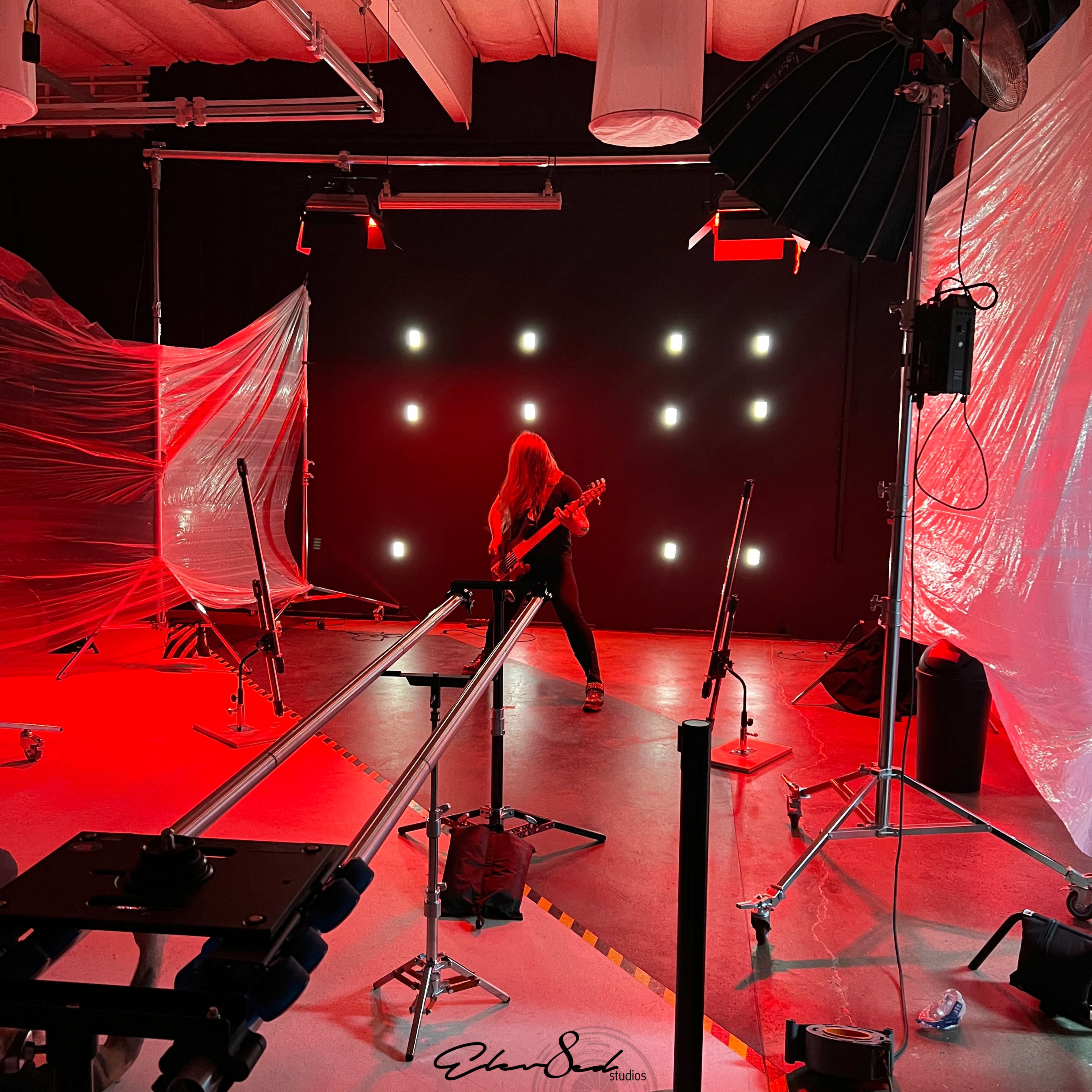 A guitarist performs on a stage surrounded by red lighting and film equipment.