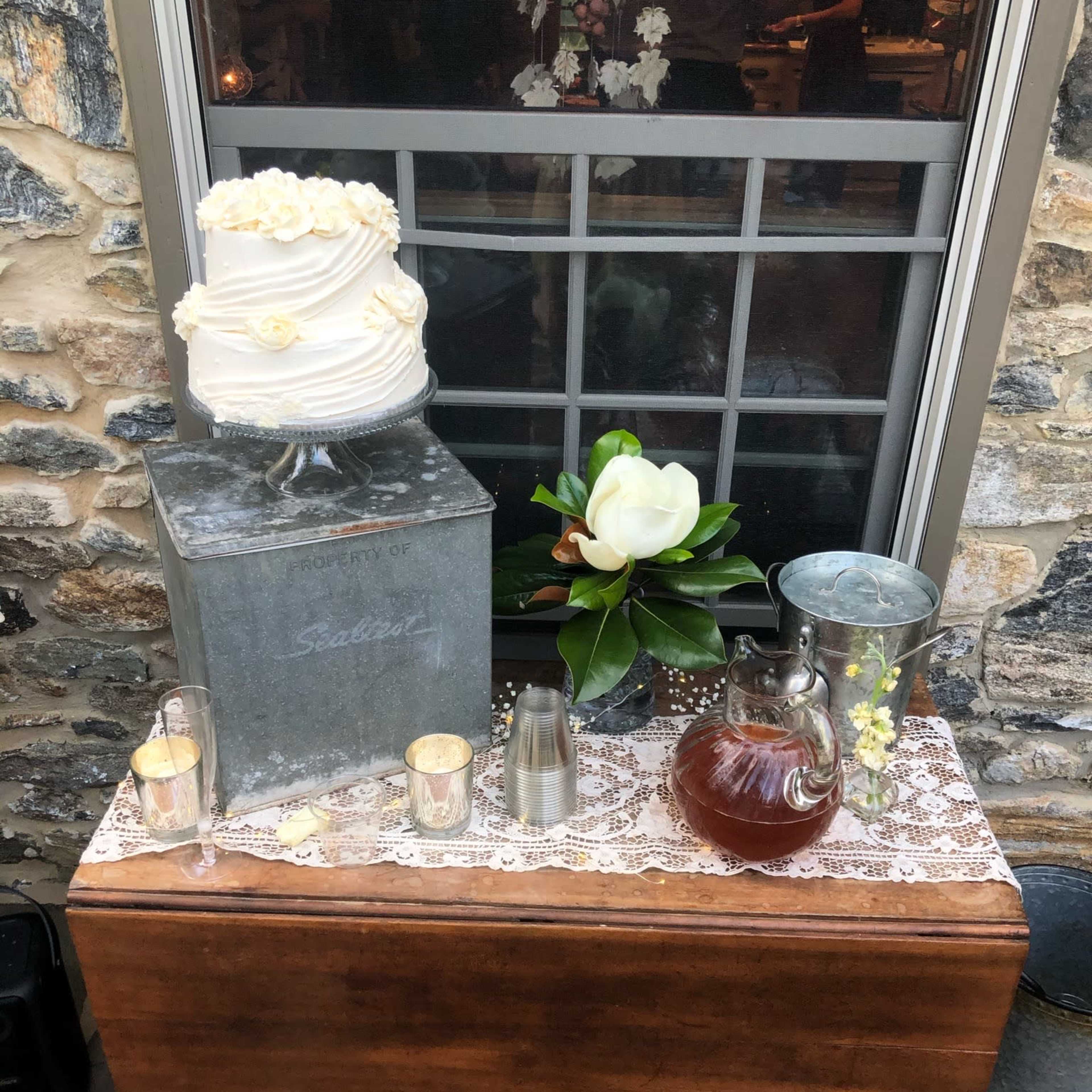 A wooden table displays a tiered wedding cake adorned with white roses, alongside a magnolia, silver pitcher, and glass jug, set against a stone wall and window.