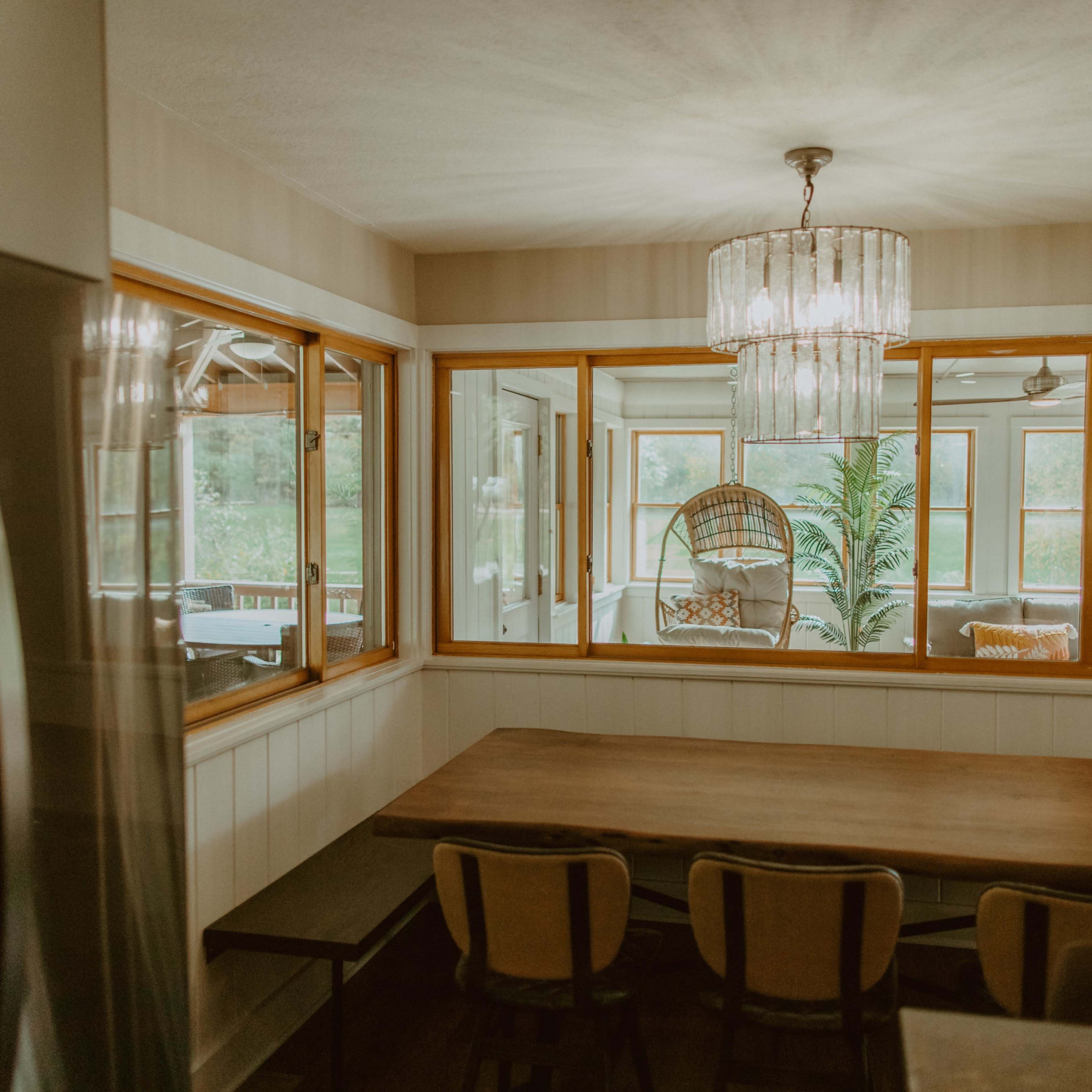 A dining area features a wooden table with chairs, adjacent to a large window that offers a view of a sunlit room with a hanging chair.