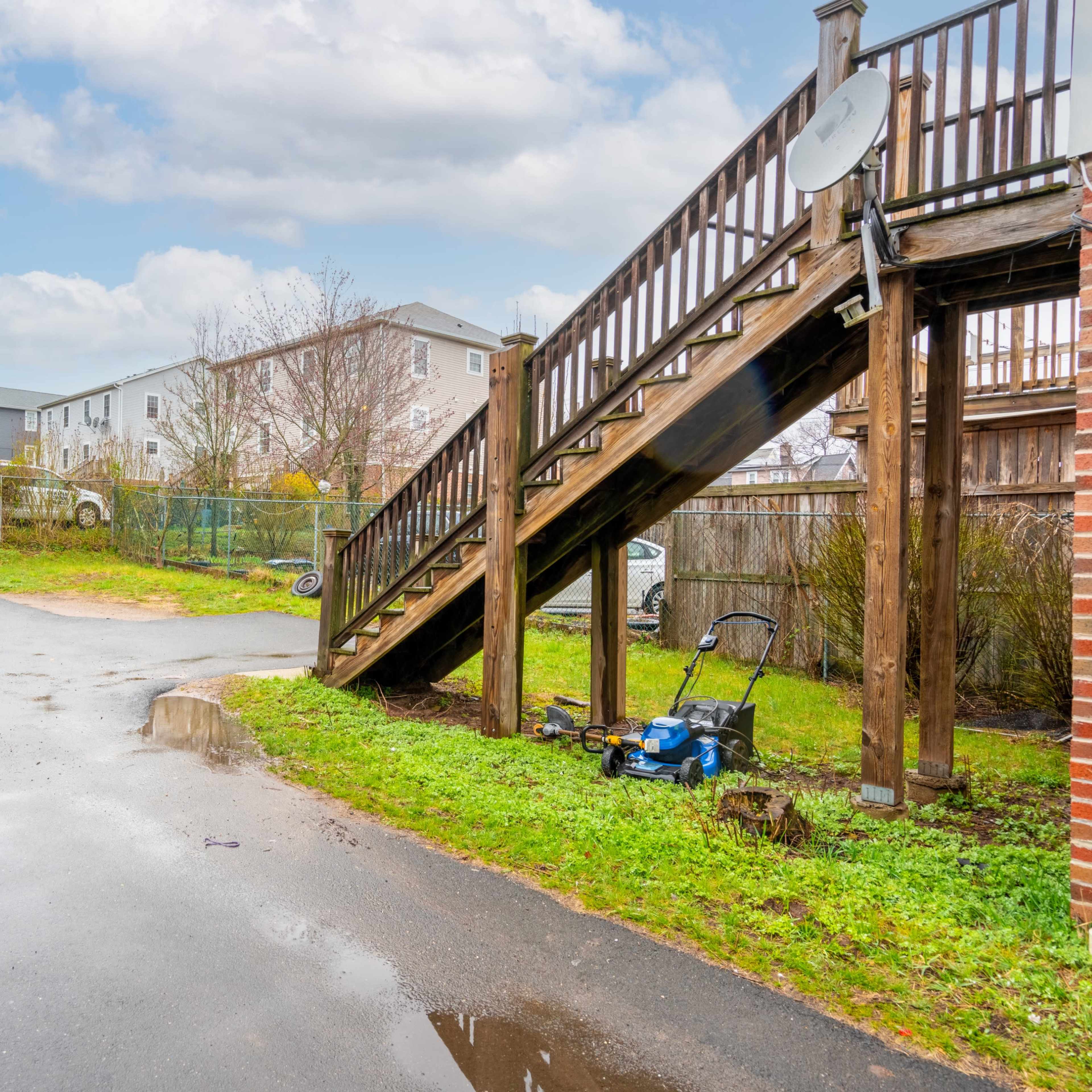 The scene features a grassy area next to a stairway leading to an upper level, with a lawnmower on the ground and several residential buildings in the background.