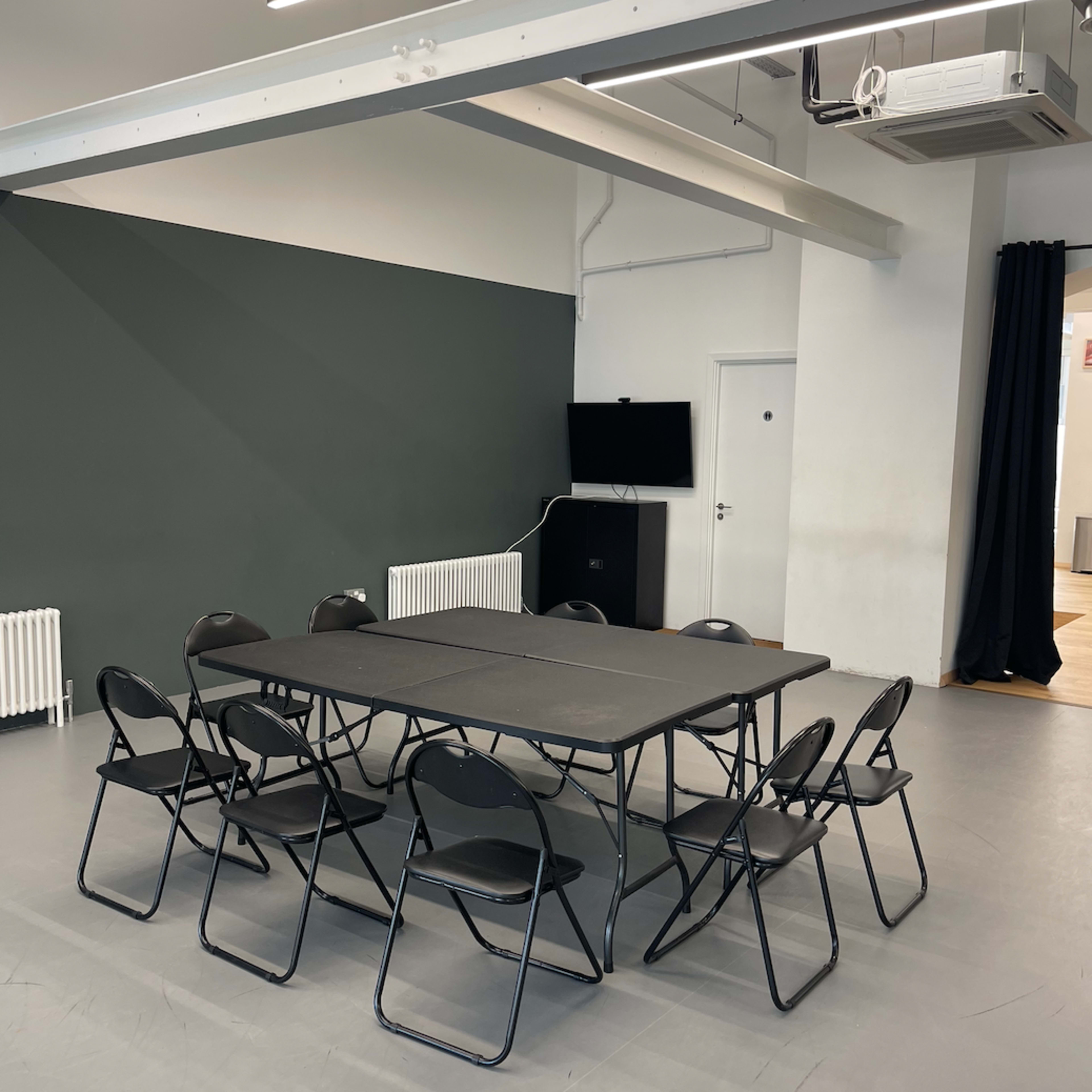 A rectangular table surrounded by eight folding chairs is set up in a minimalist room with a dark green wall and a door.