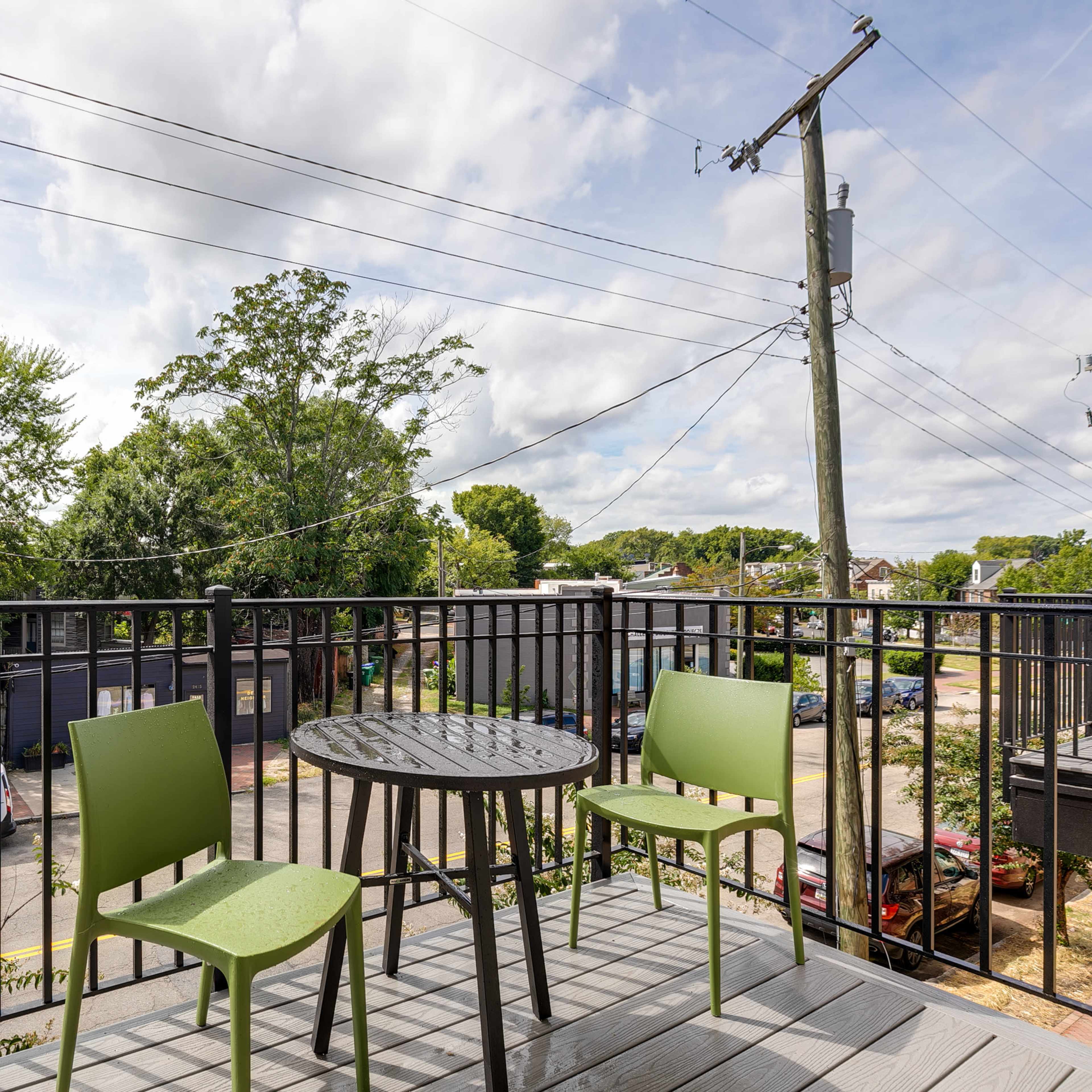 A balcony with two green chairs and a small round table overlooking a tree-lined street with power lines in the background.
