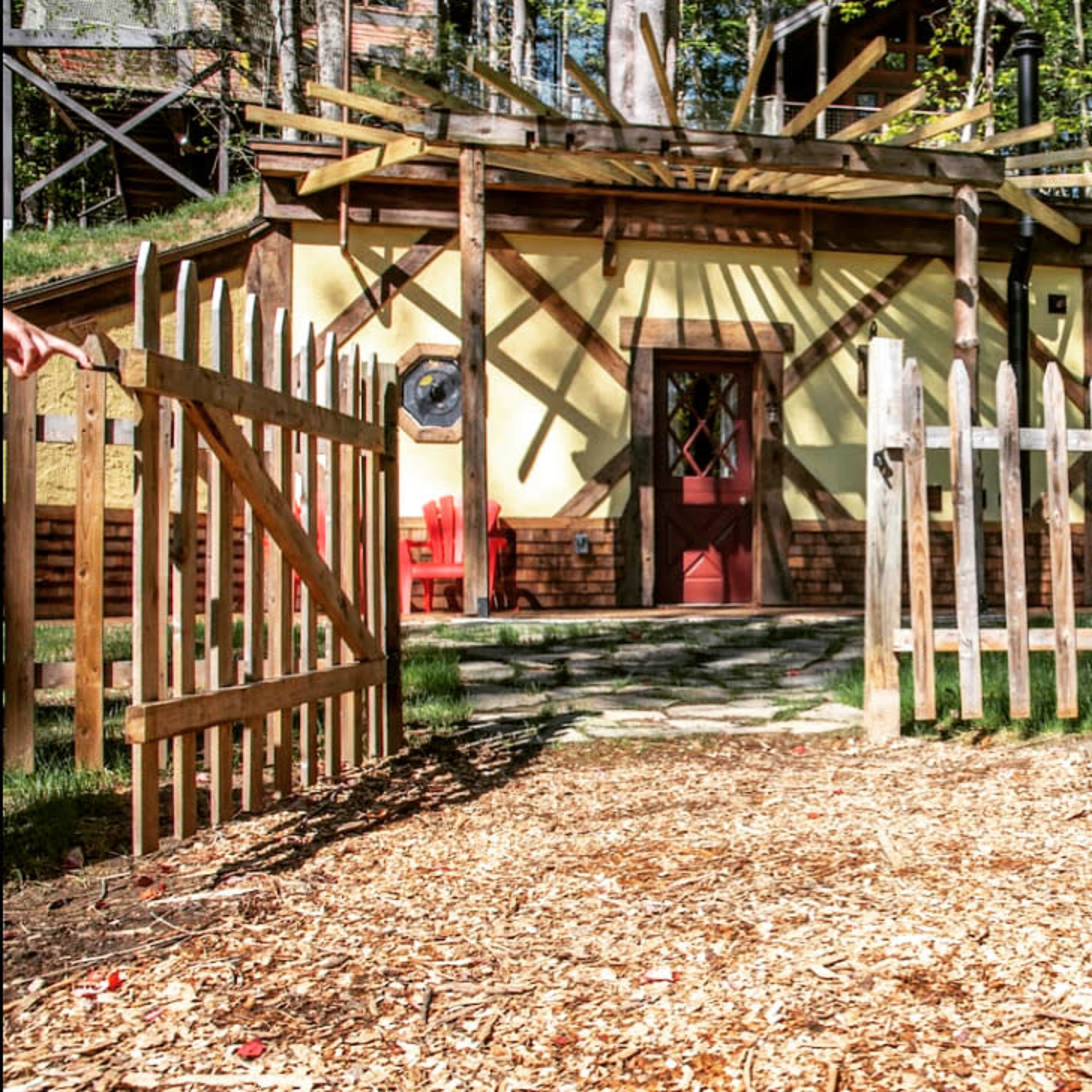A wooden fence leads to a house nestled among trees, with a red door and a pathway made of stones and mulch.