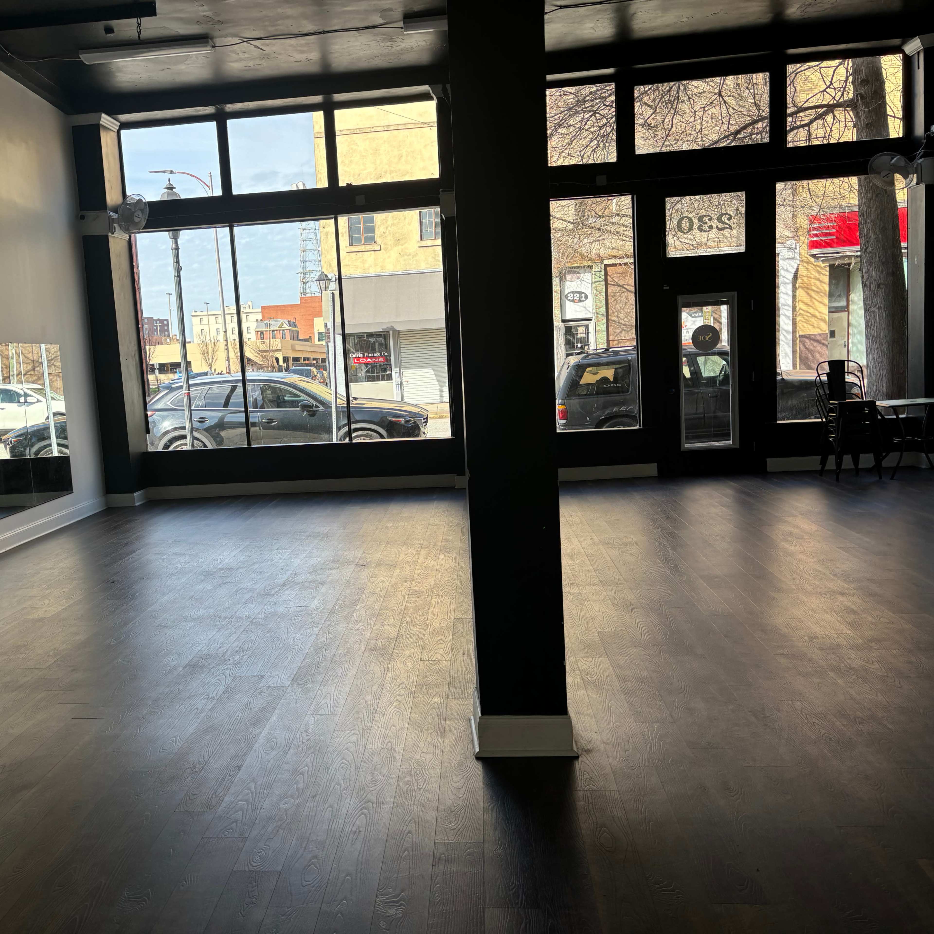 A vacant interior space with wooden flooring and large windows facing a street.